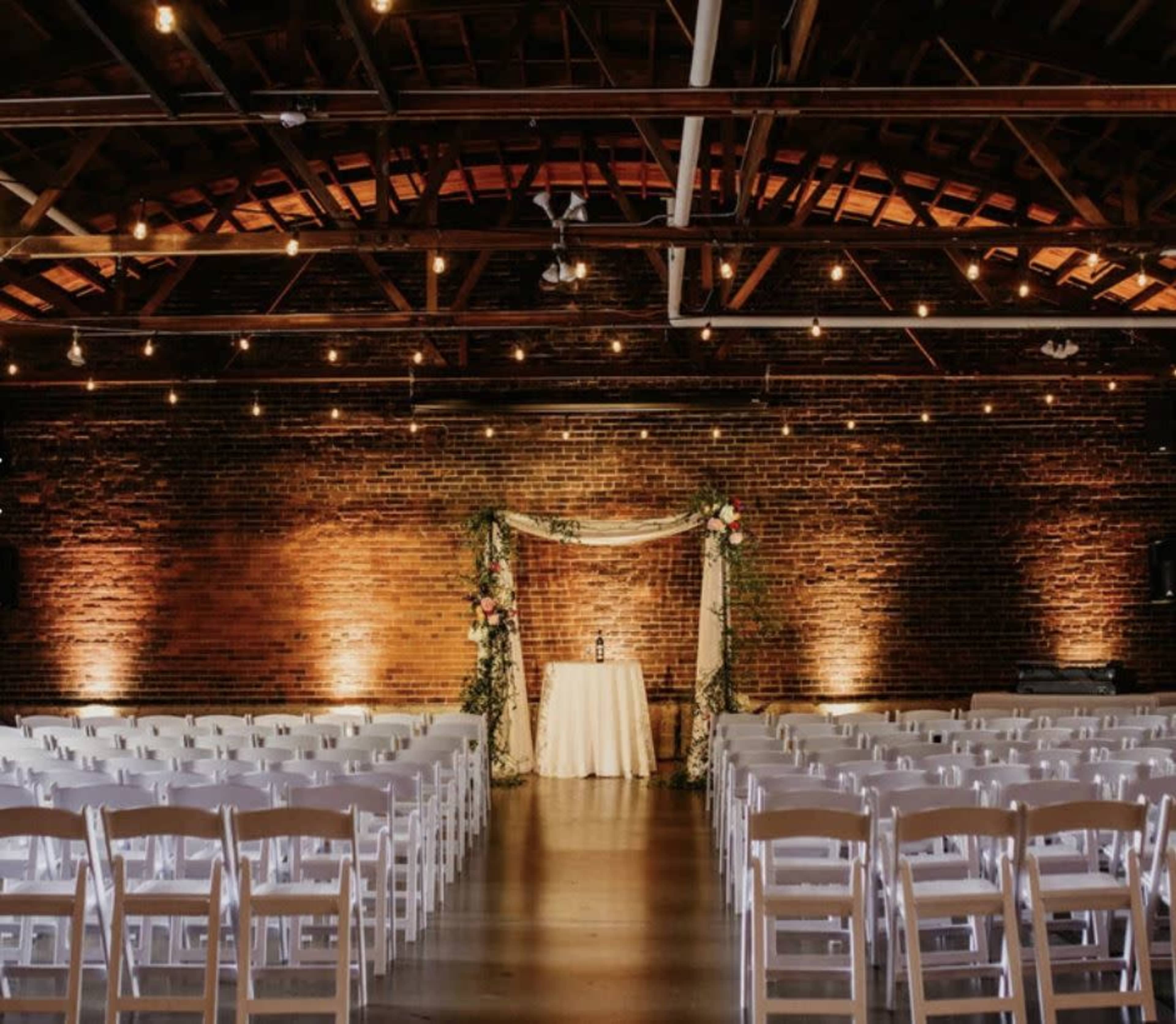 A wedding ceremony setup features rows of white chairs facing a decorated altar against a brick wall in a softly lit venue.
