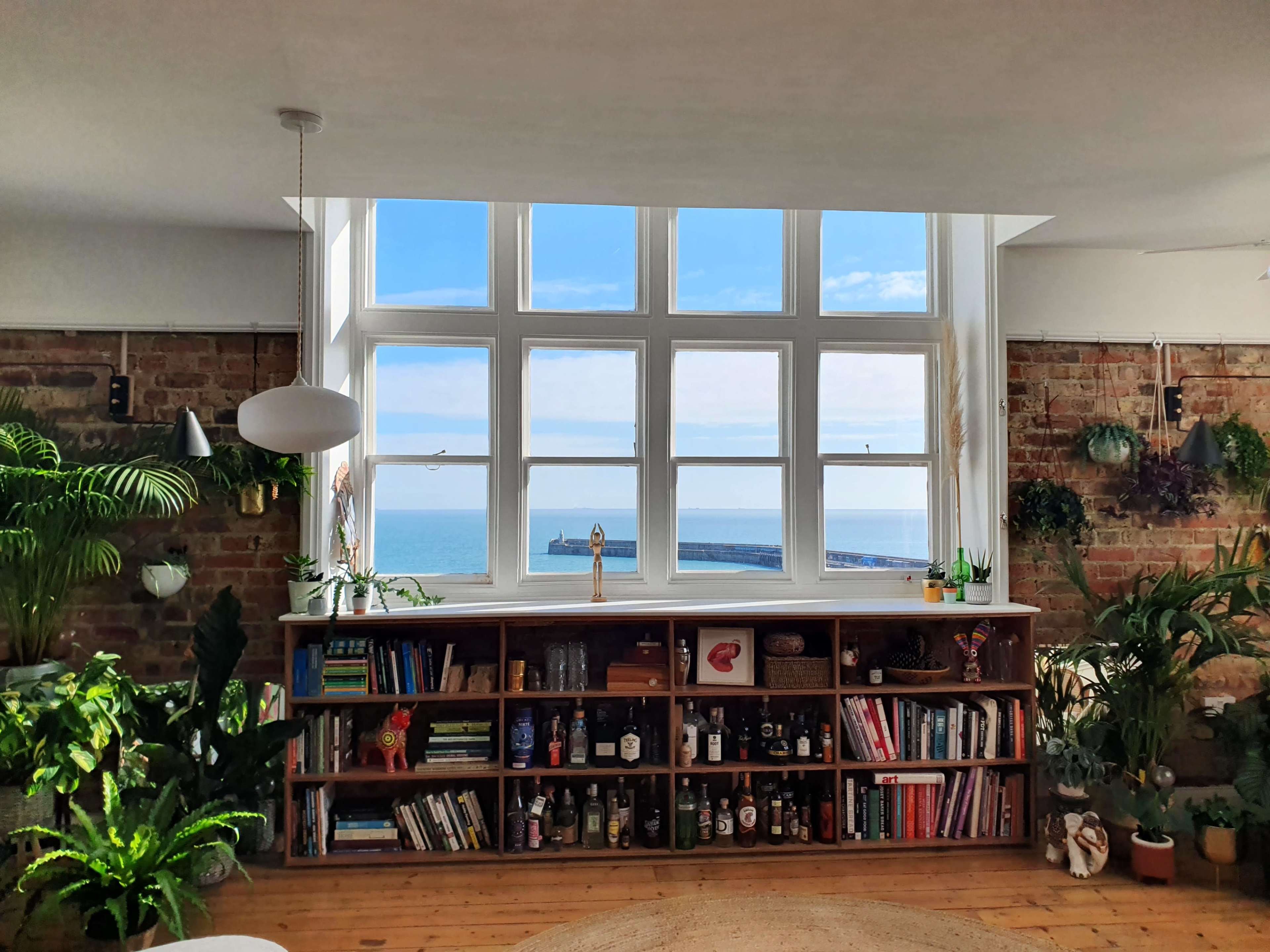 A spacious room with a large window overlooking the sea, featuring a bookshelf filled with various books and plants.