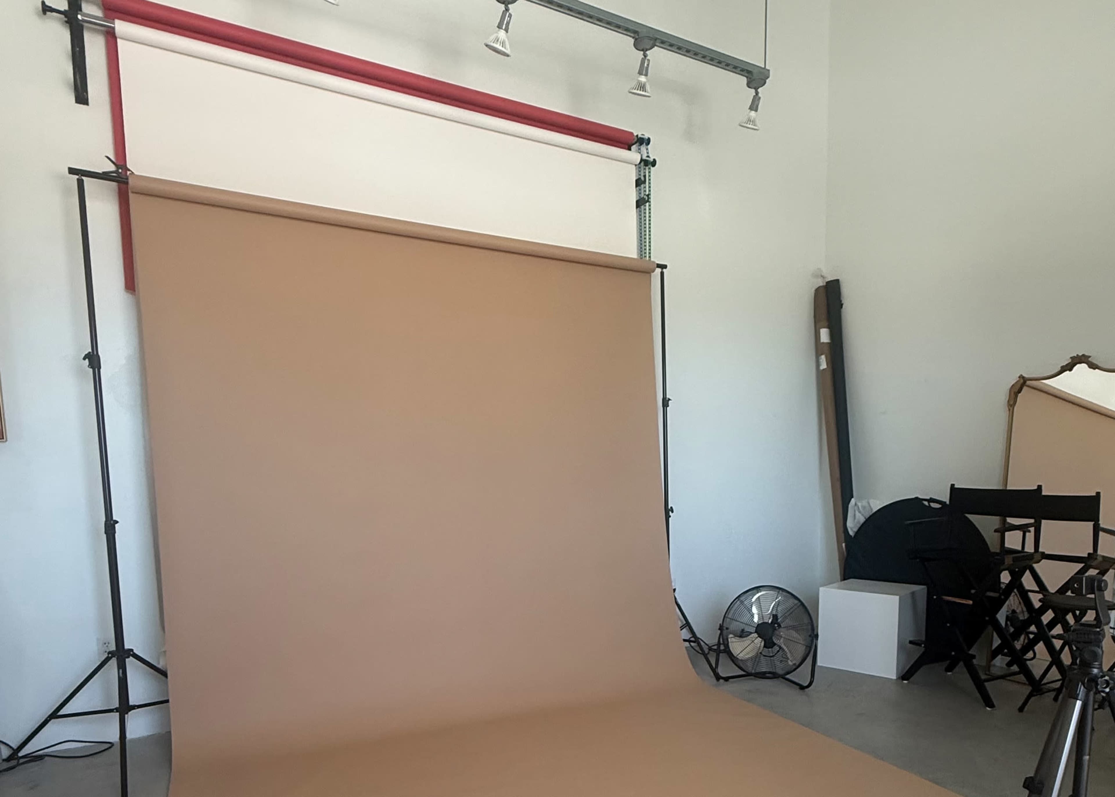 The image shows a photography studio with a neutral-colored backdrop, lighting equipment, and a few studio chairs.