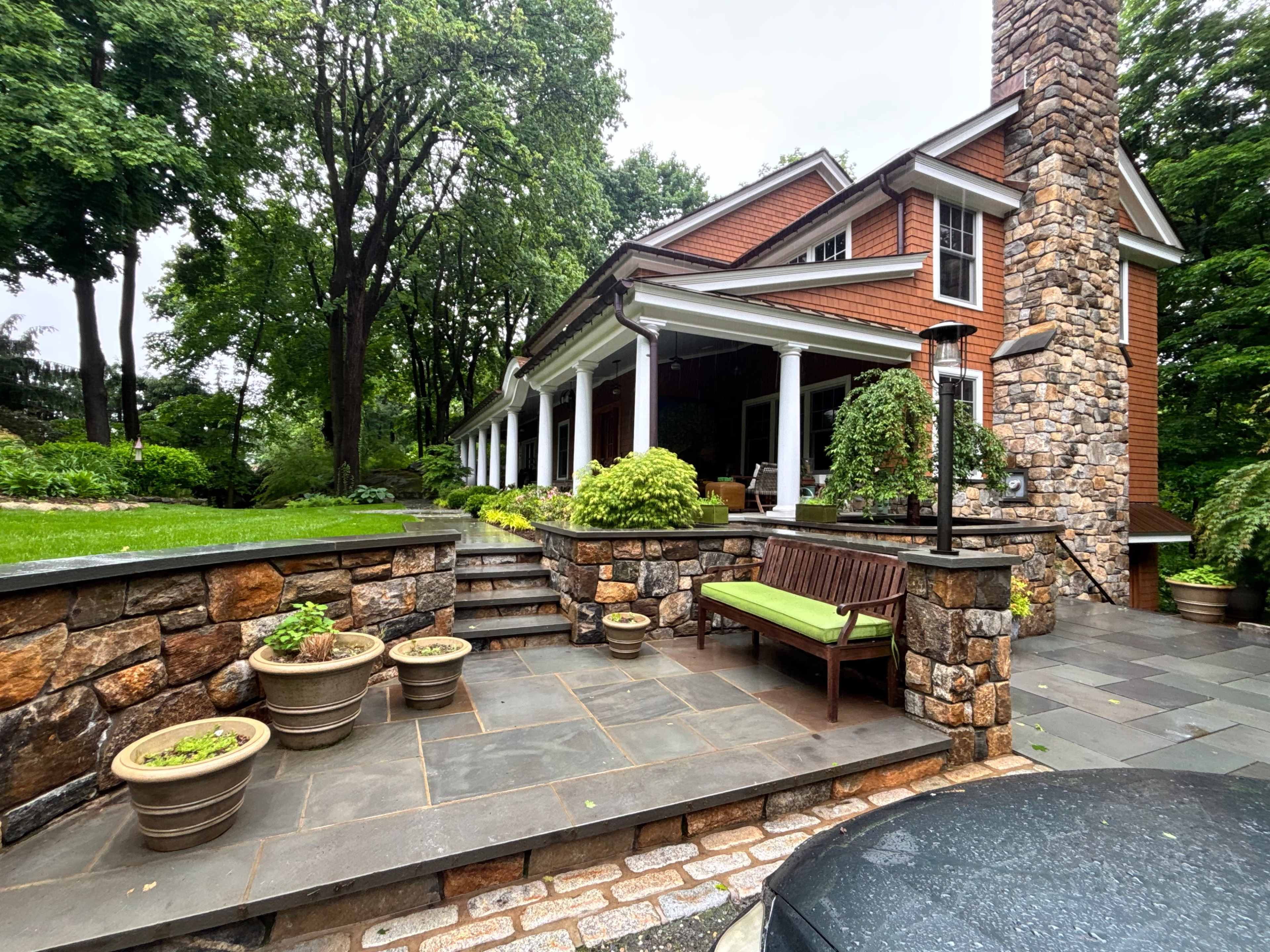 A two-story brown wooden house with a stone chimney is surrounded by green trees and features a patio with stone steps and potted plants.
