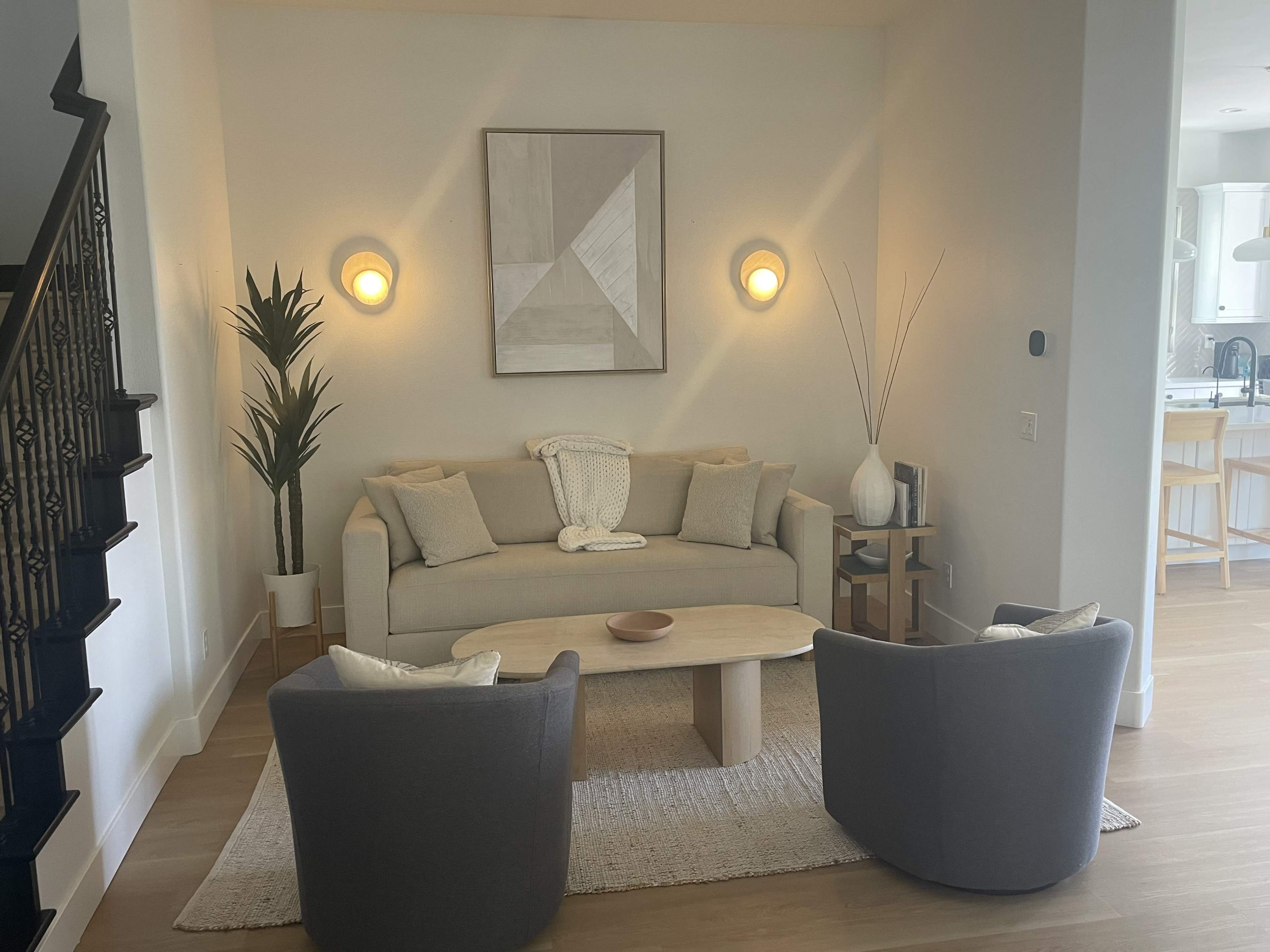 A light-filled living room features a gray sofa, two matching chairs, a round coffee table, and wall-mounted lights against a neutral backdrop.