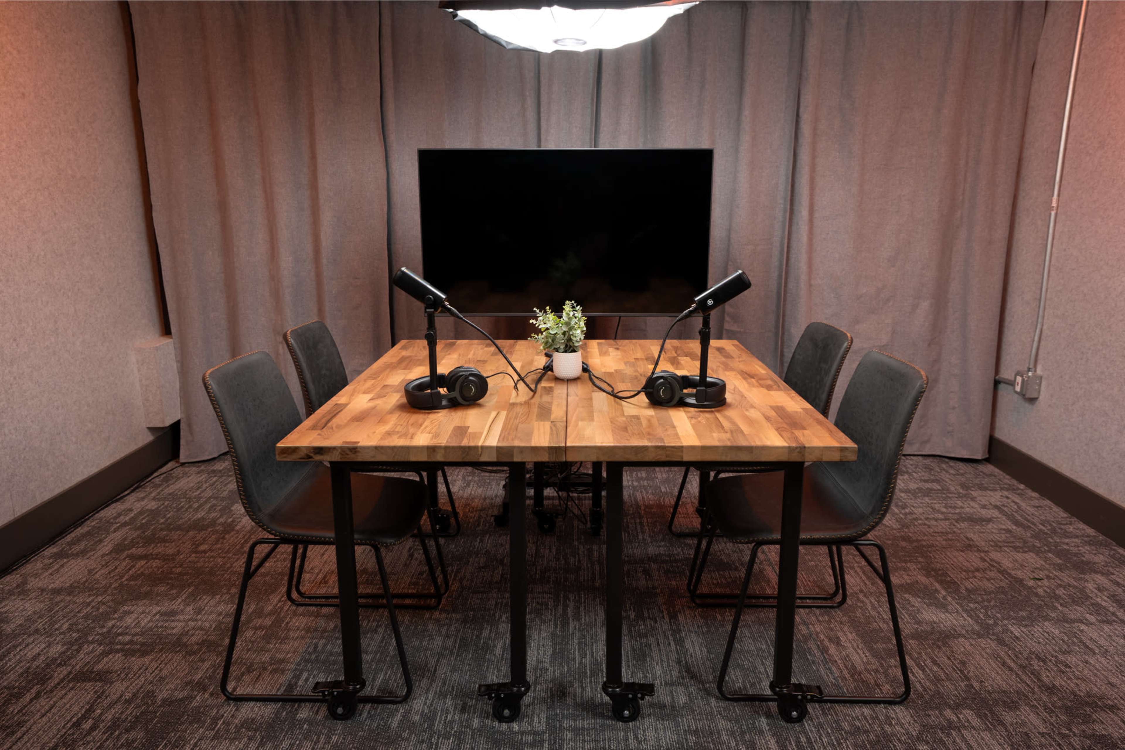 A rectangular wooden table with four chairs and headsets is positioned in front of a large screen, set against a backdrop of gray curtains.