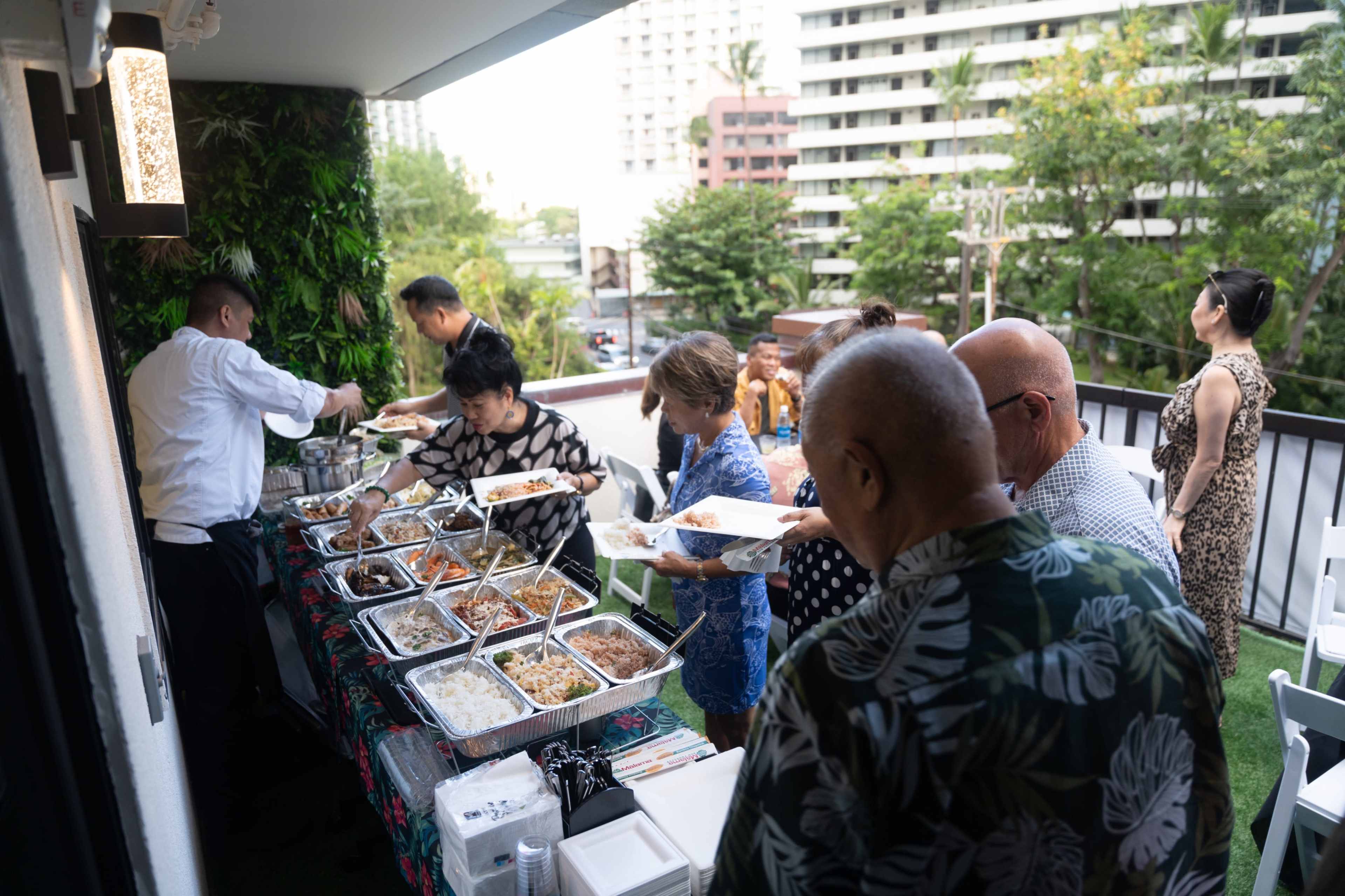 A group of people is serving themselves from a buffet spread on a balcony with a view of urban greenery.