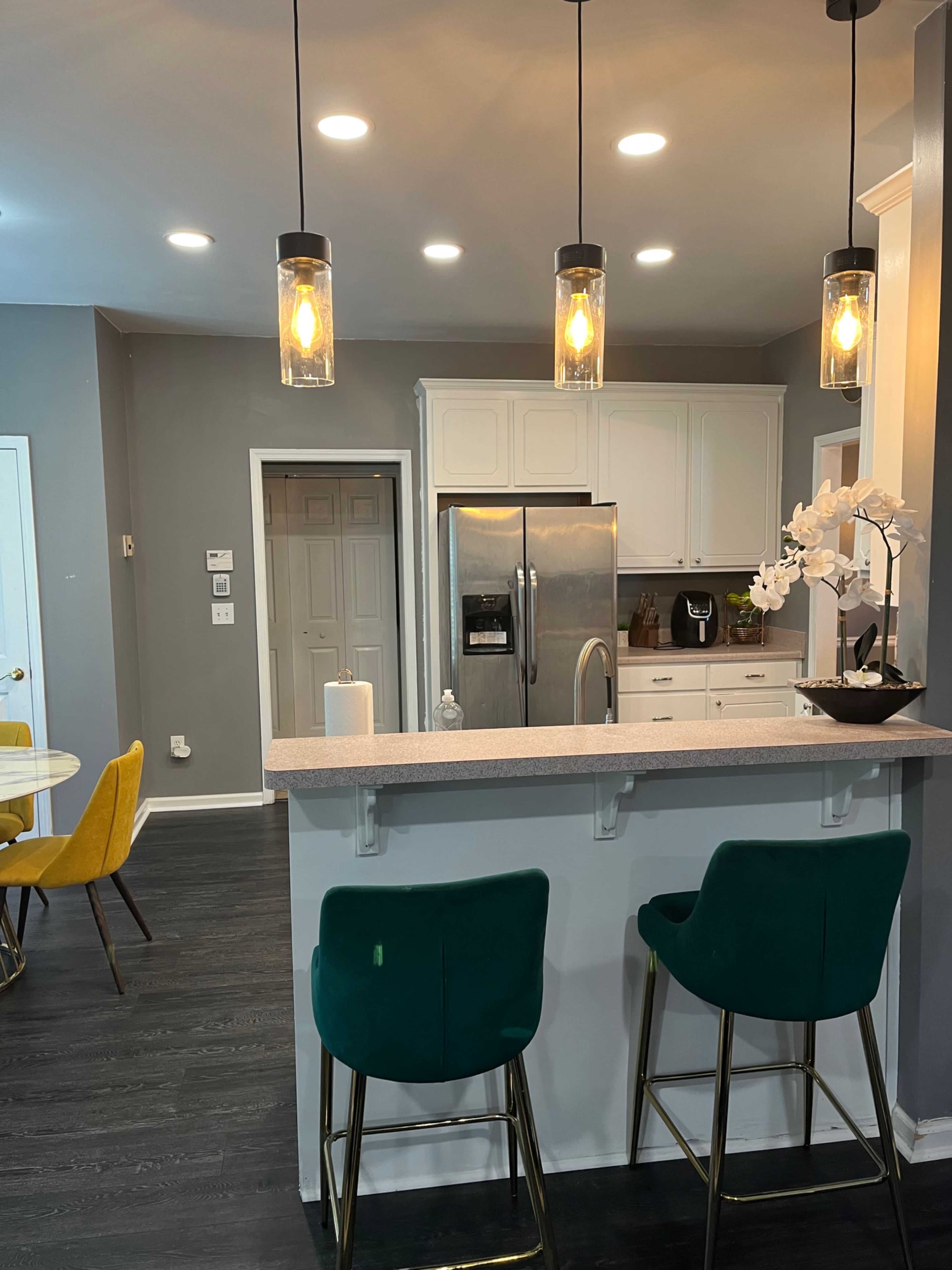 The image shows a modern kitchen with a bar area featuring two green stools, pendant lights, and white cabinets.