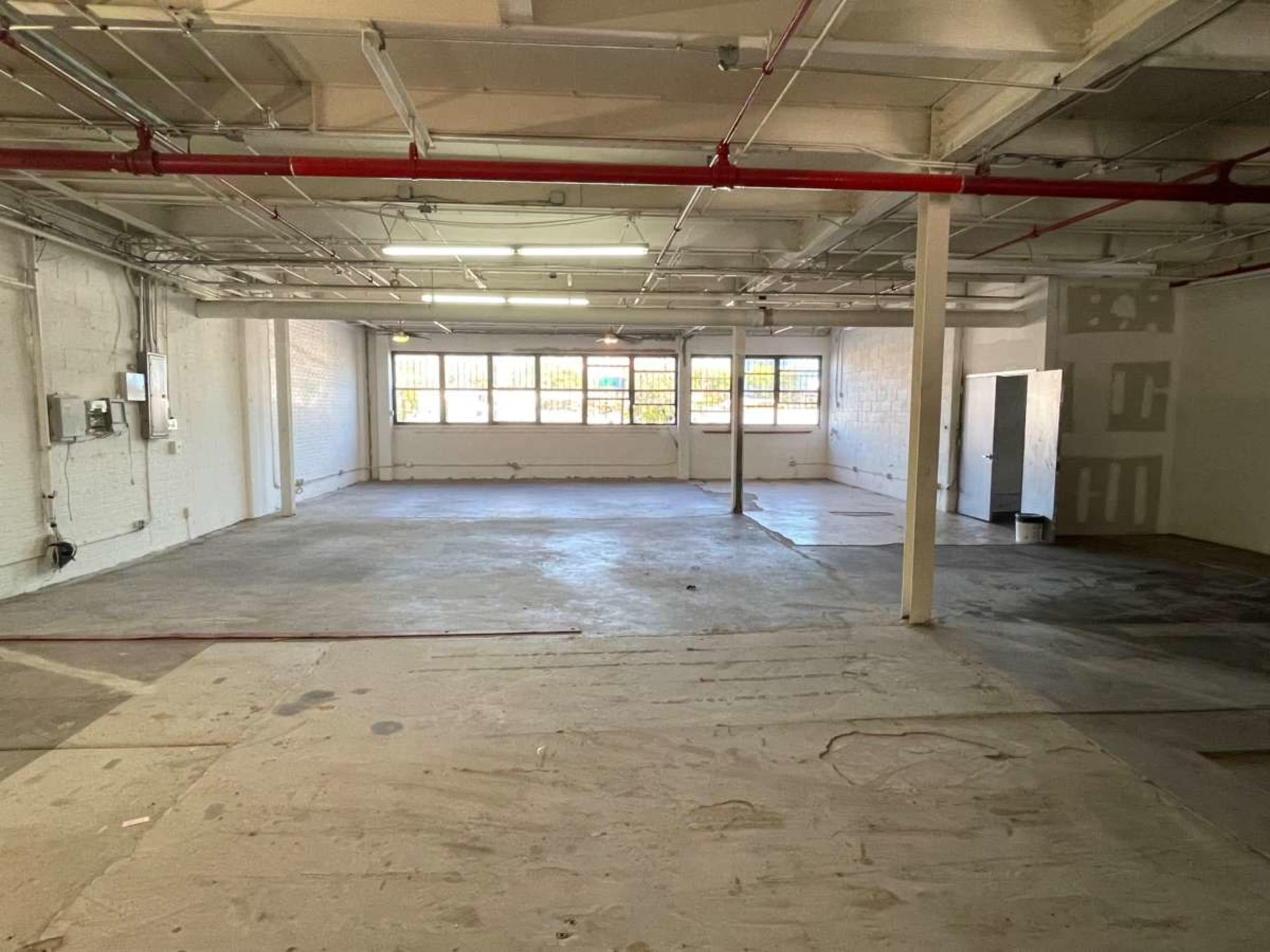 The image shows a large, empty industrial space with exposed ceiling beams and natural light coming through multiple windows.