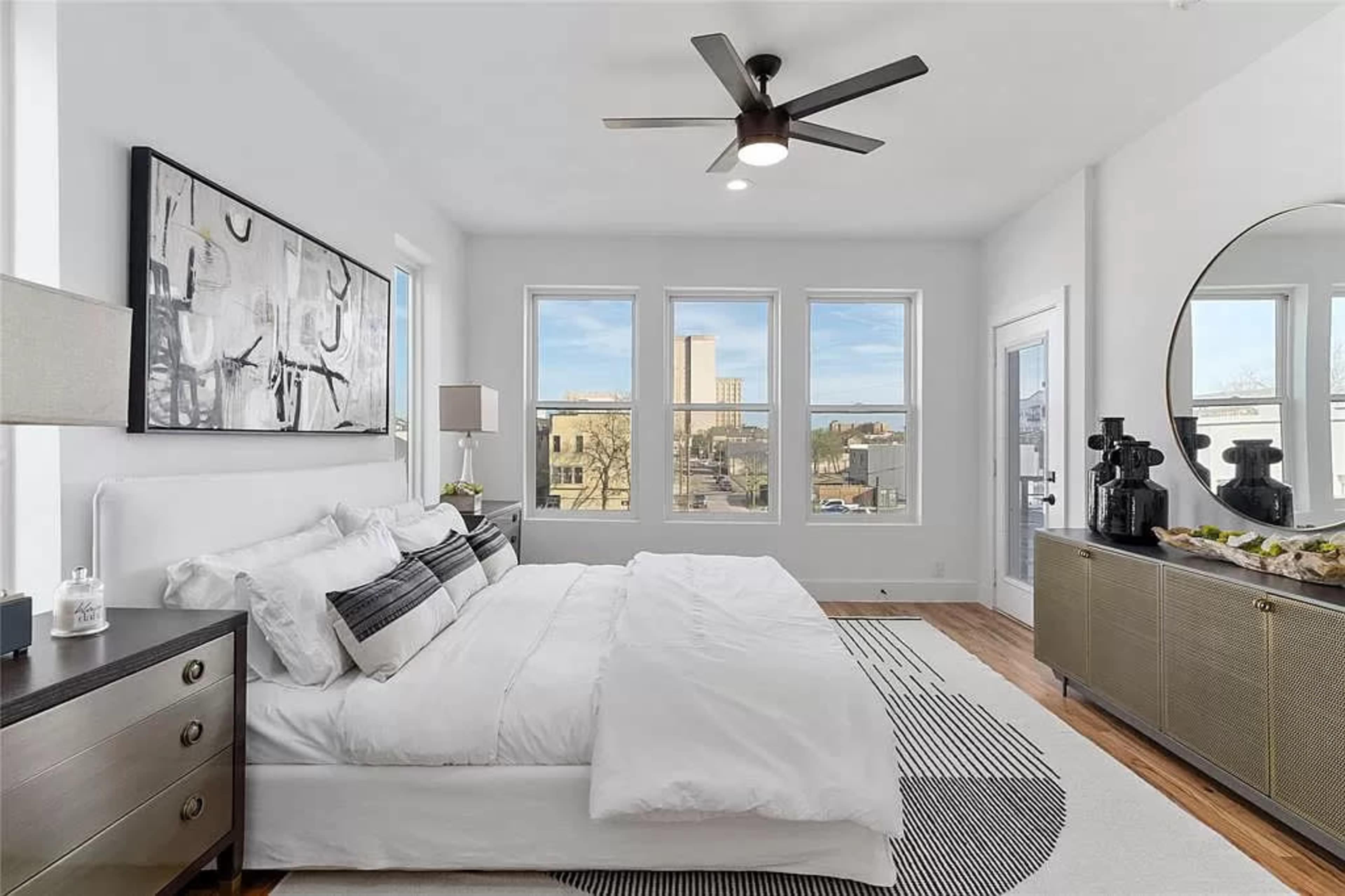 The bedroom features a large bed with white bedding, three windows providing natural light, and modern furniture, including a sleek dresser and a round mirror.