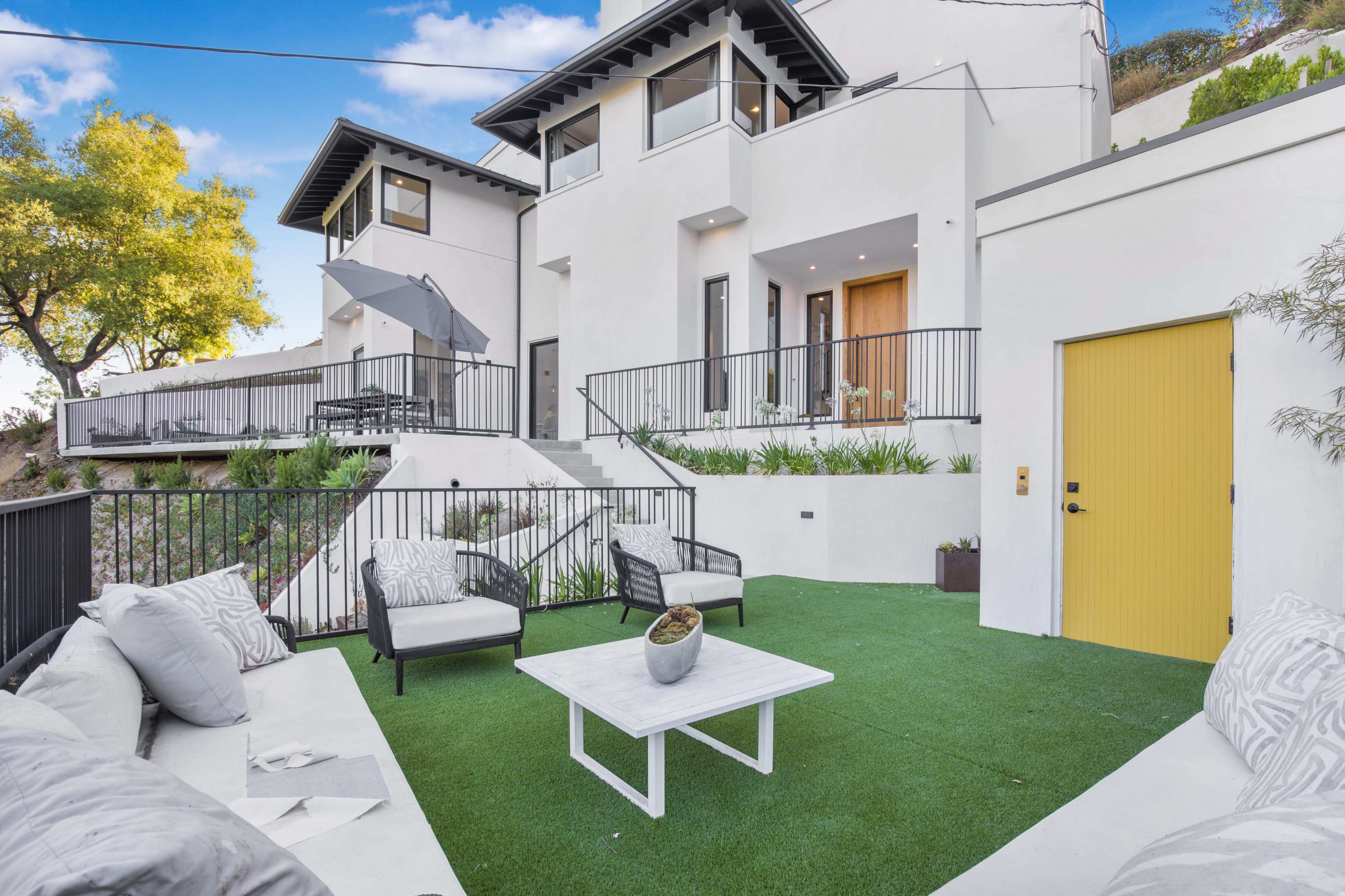 The image shows a modern outdoor living space featuring a white sofa set with cushions, a coffee table, and a yellow door on a hillside property.