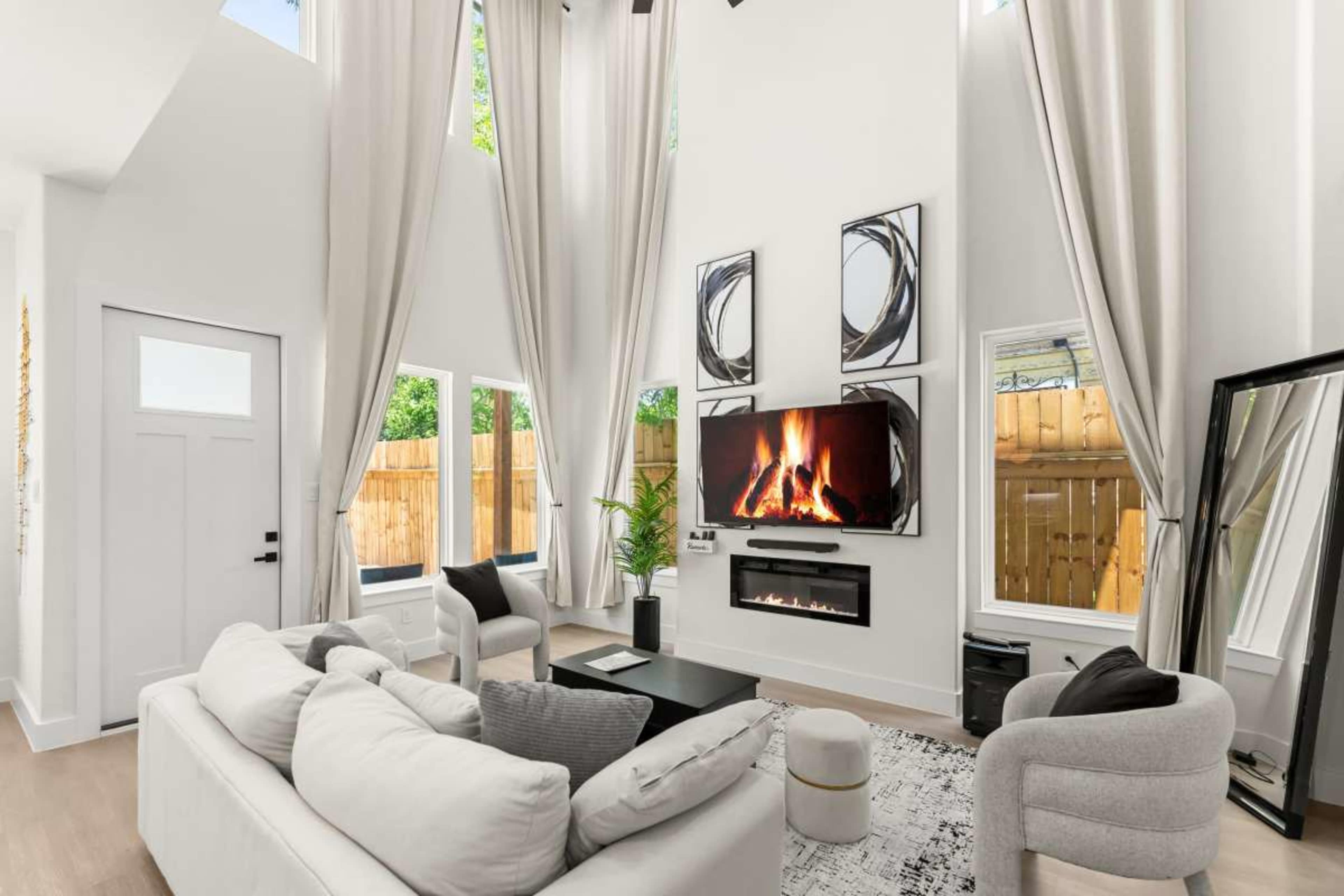 The scene features a modern living room with high ceilings, large windows, a fireplace beneath a mounted TV, and contemporary furniture.