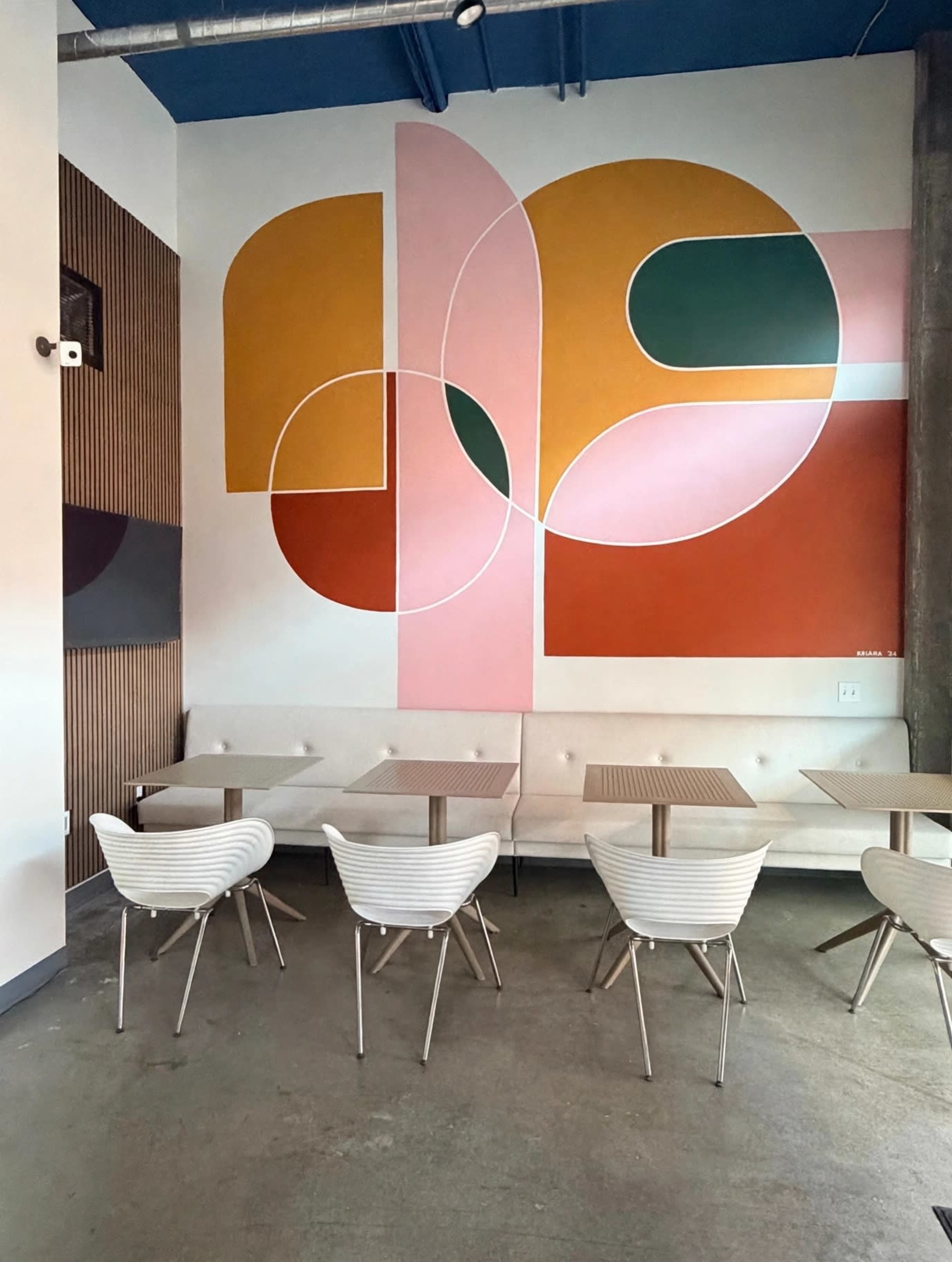 A seating area in a café features white furniture and a large colorful abstract mural on the wall.