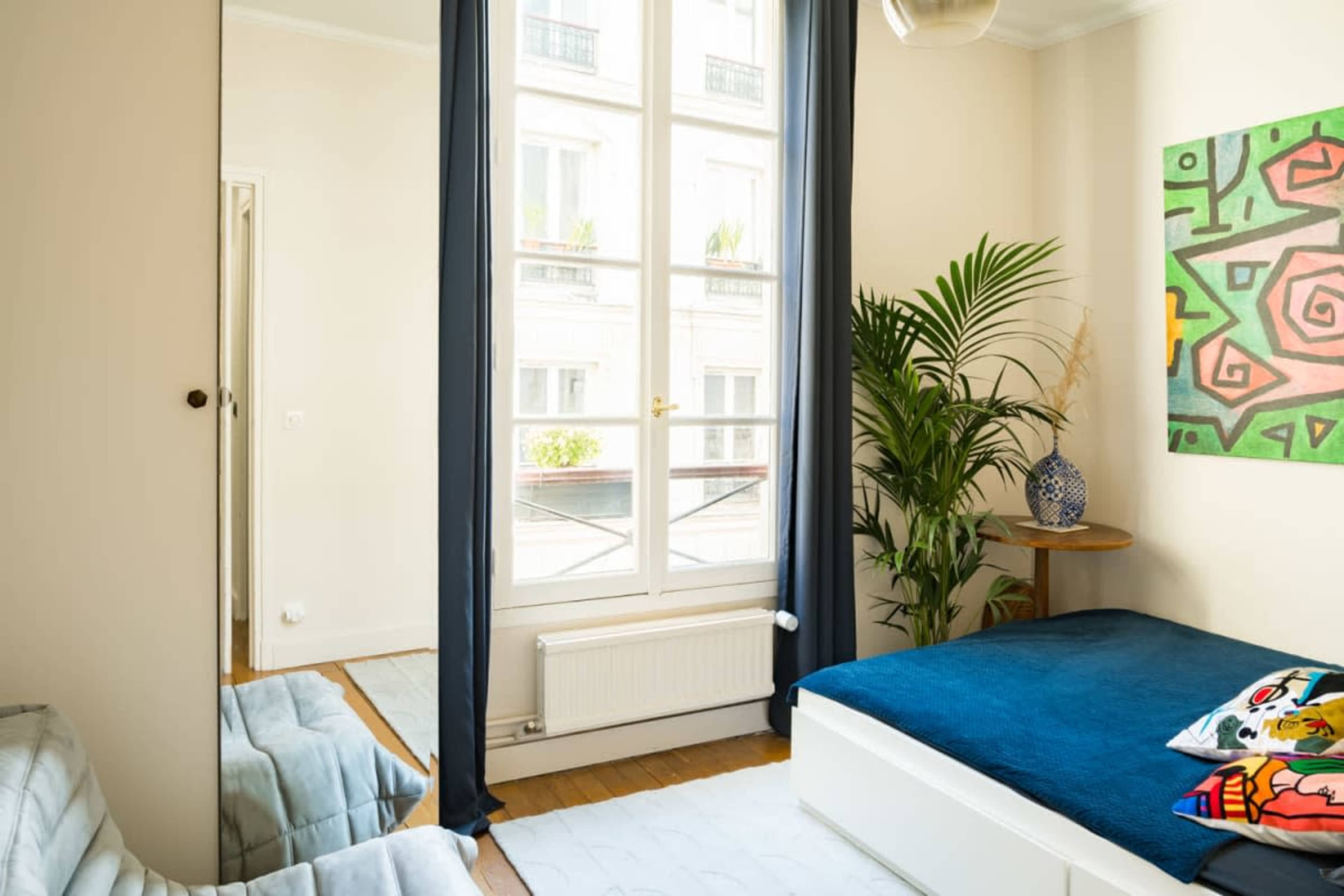 A bedroom with a blue bedspread, a potted plant, and a colorful abstract painting on the wall, illuminated by natural light from a large window.