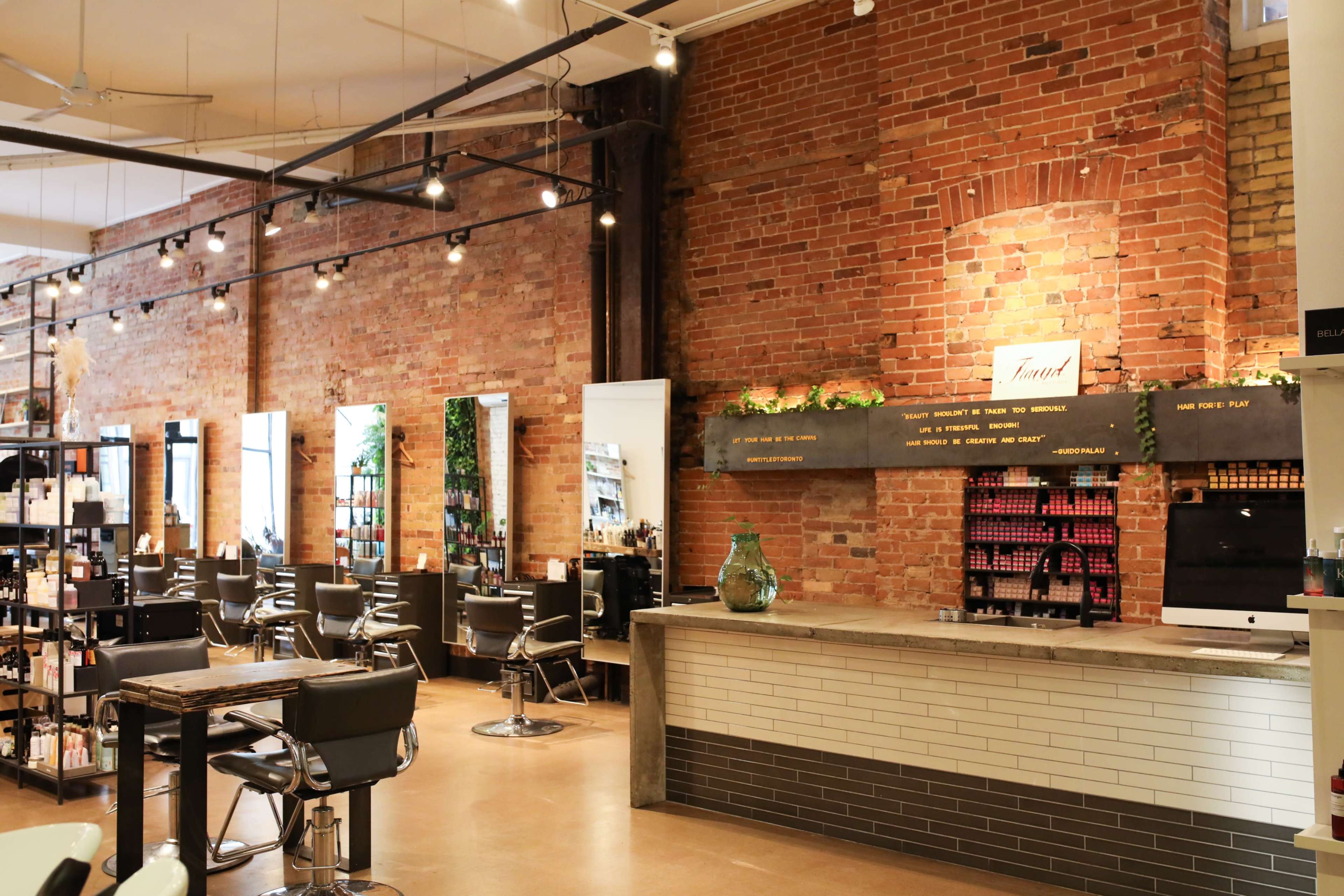 The interior of a modern hair salon with brick walls, styling stations, and a reception area featuring decorative shelves and a computer.