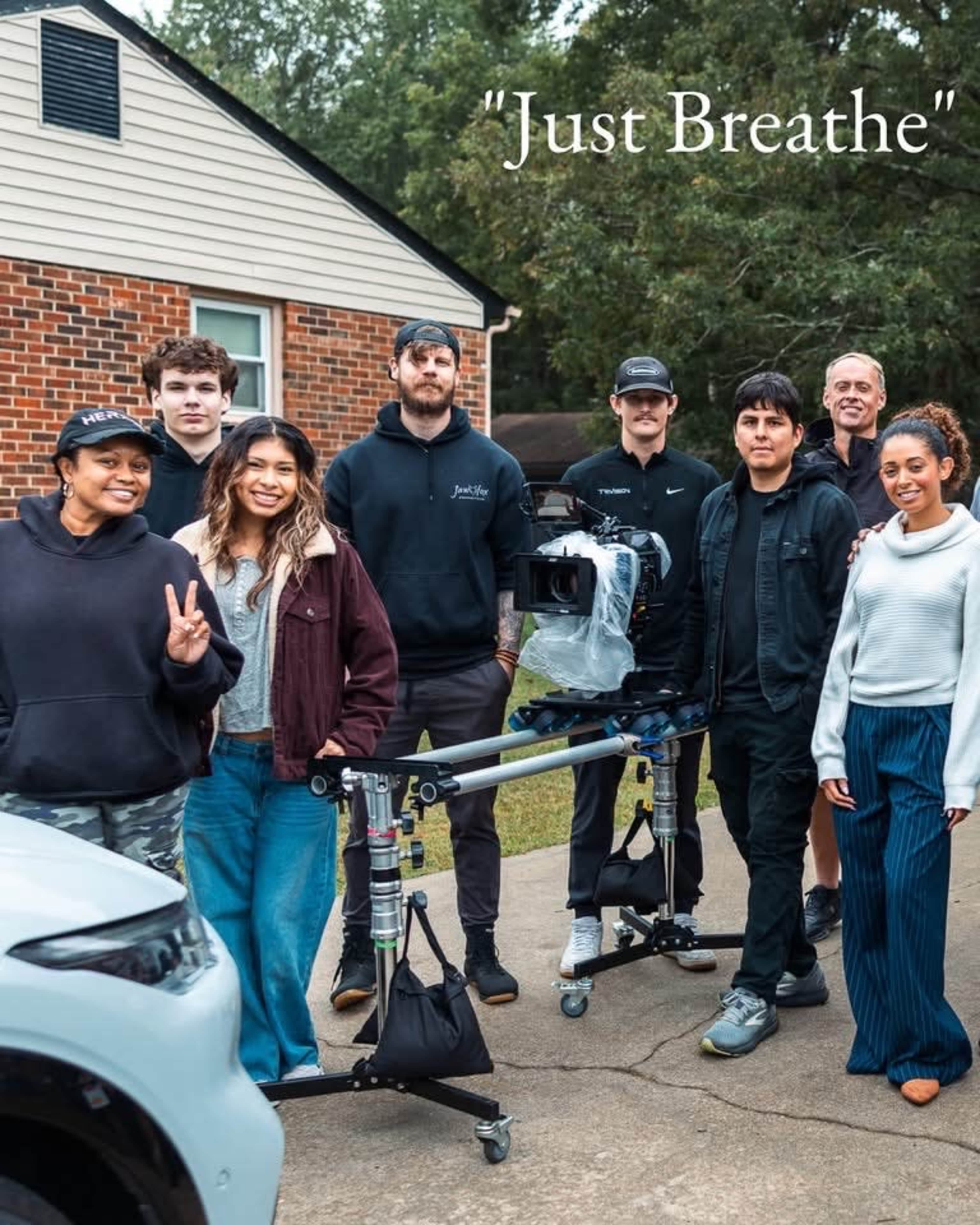A group of nine people poses together outdoors near a car and a house, with a camera on a tripod in the foreground.