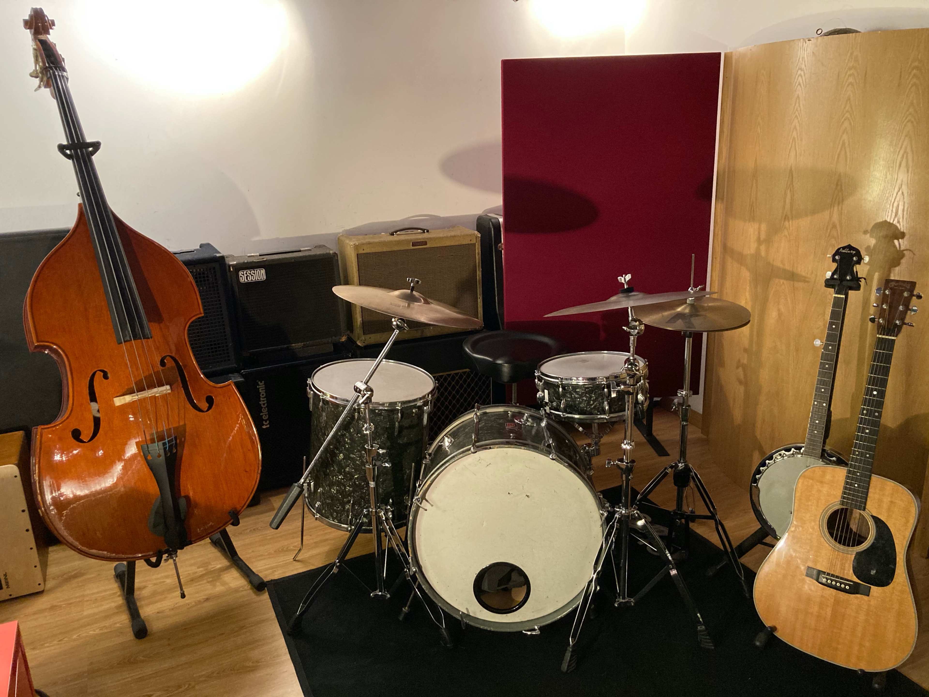 The scene features a double bass, a drum set, and an acoustic guitar arranged in a music rehearsal space surrounded by amplifiers.