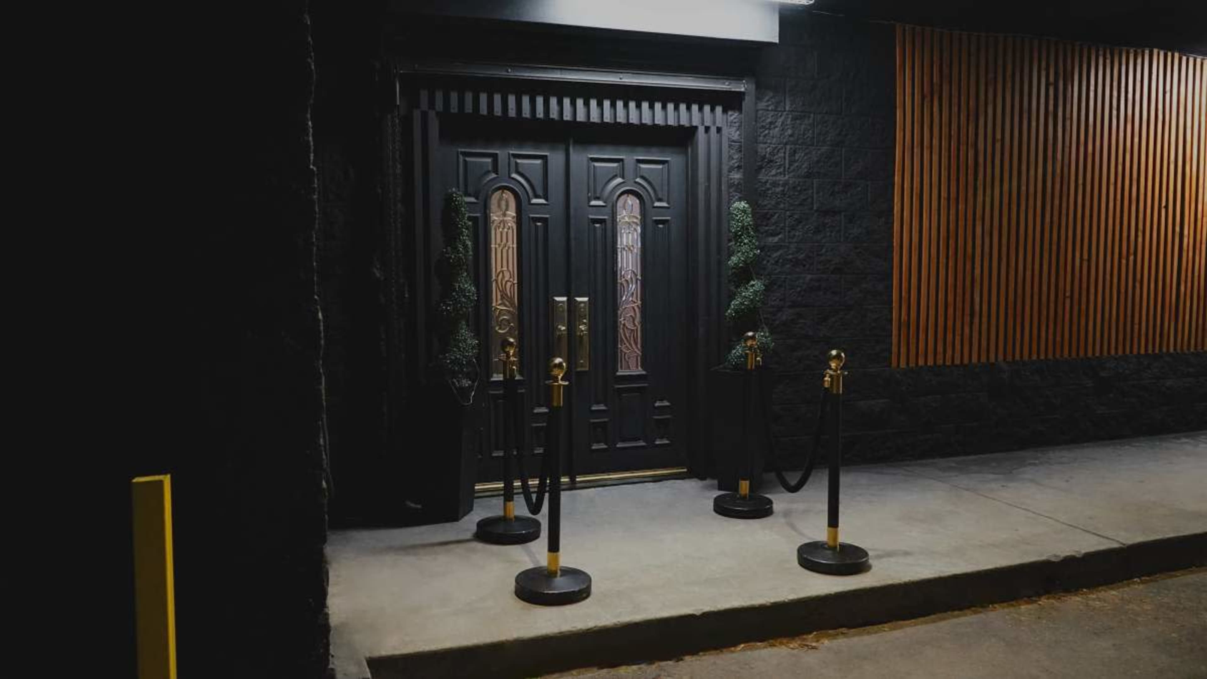 The image shows a dark entrance with a black door featuring decorative elements, flanked by ropes and stanchions on the ground.