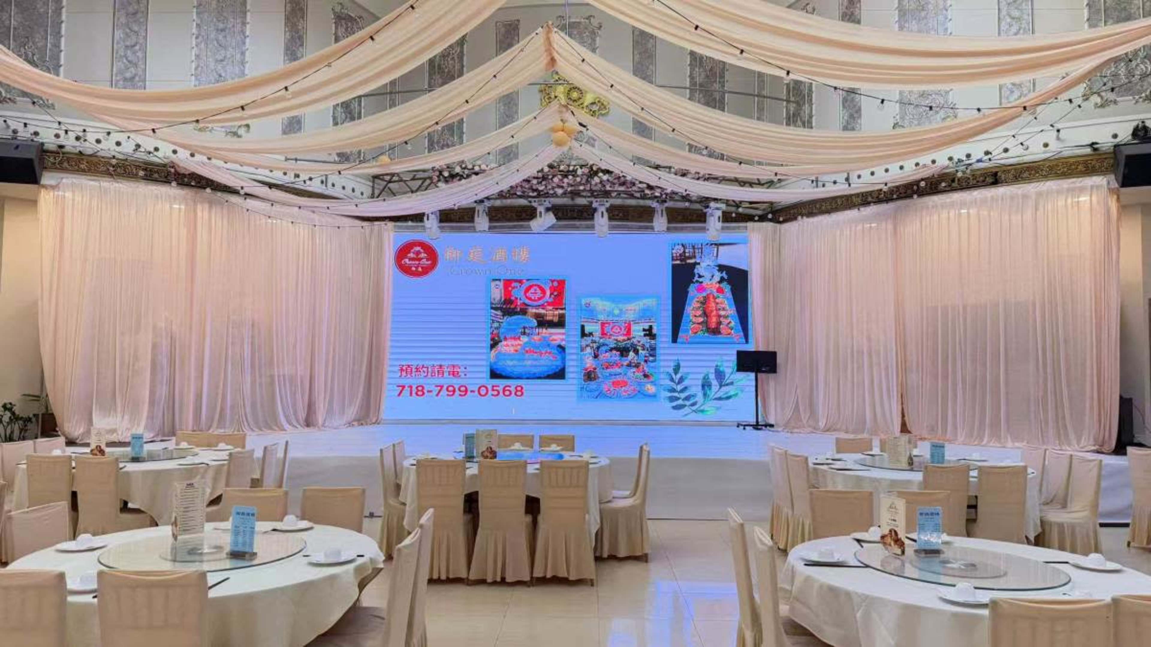 The image shows an elegantly arranged banquet hall with round tables, a large screen displaying promotional content, and draped fabric overhead.