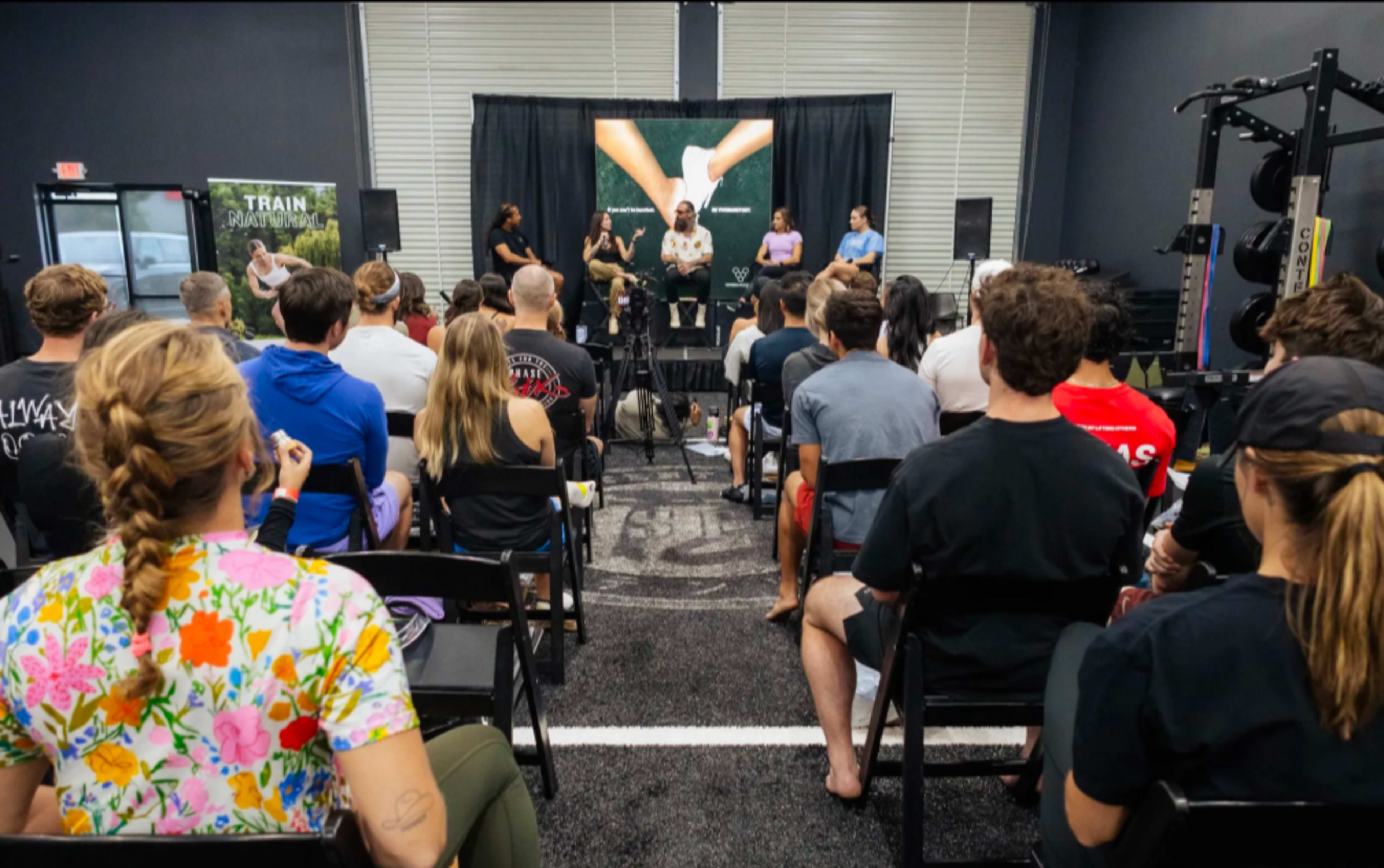 A group of people is seated in a fitness facility, listening to a panel discussion on stage.