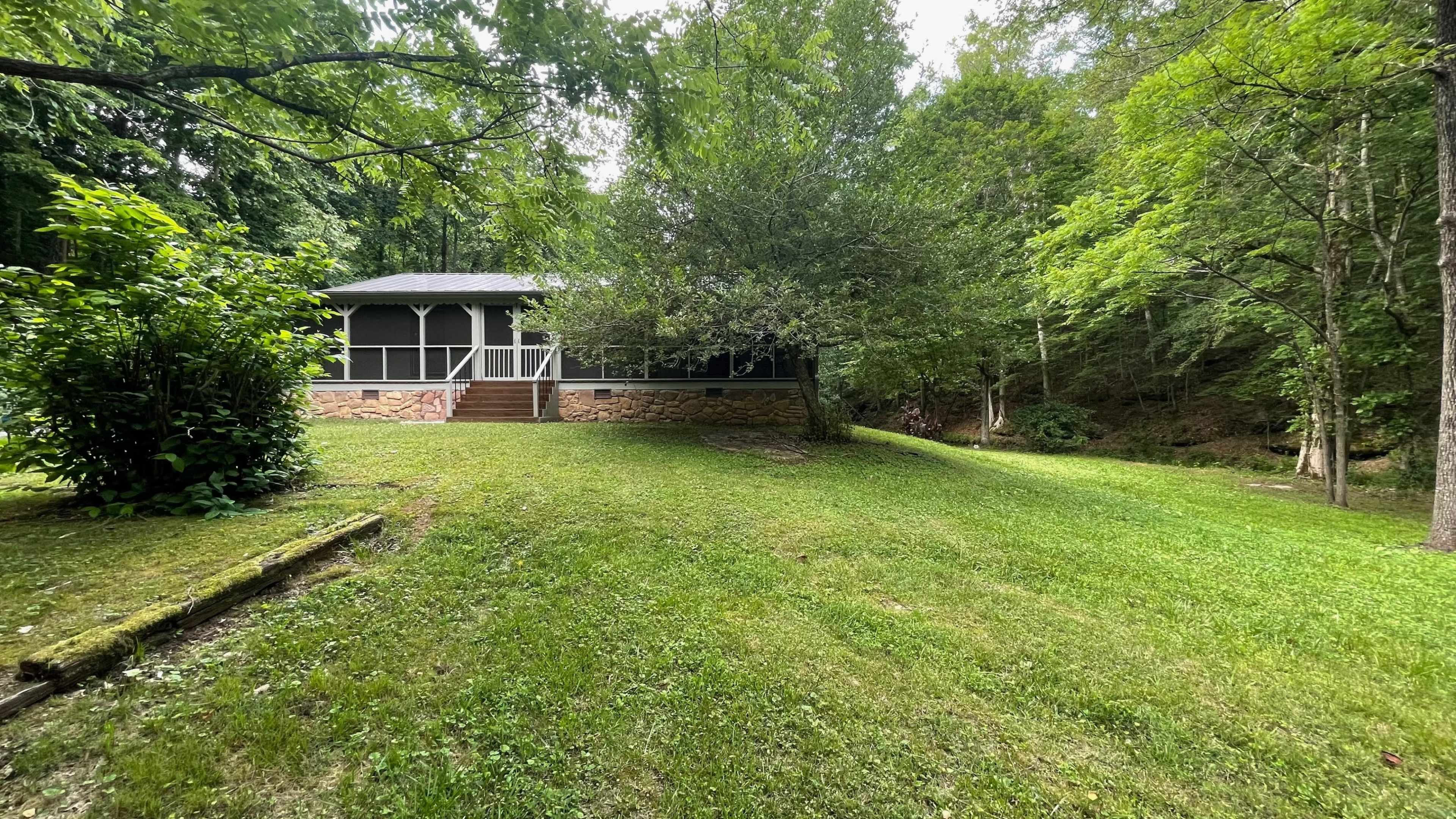 A screened porch house is situated on a grassy slope surrounded by trees.