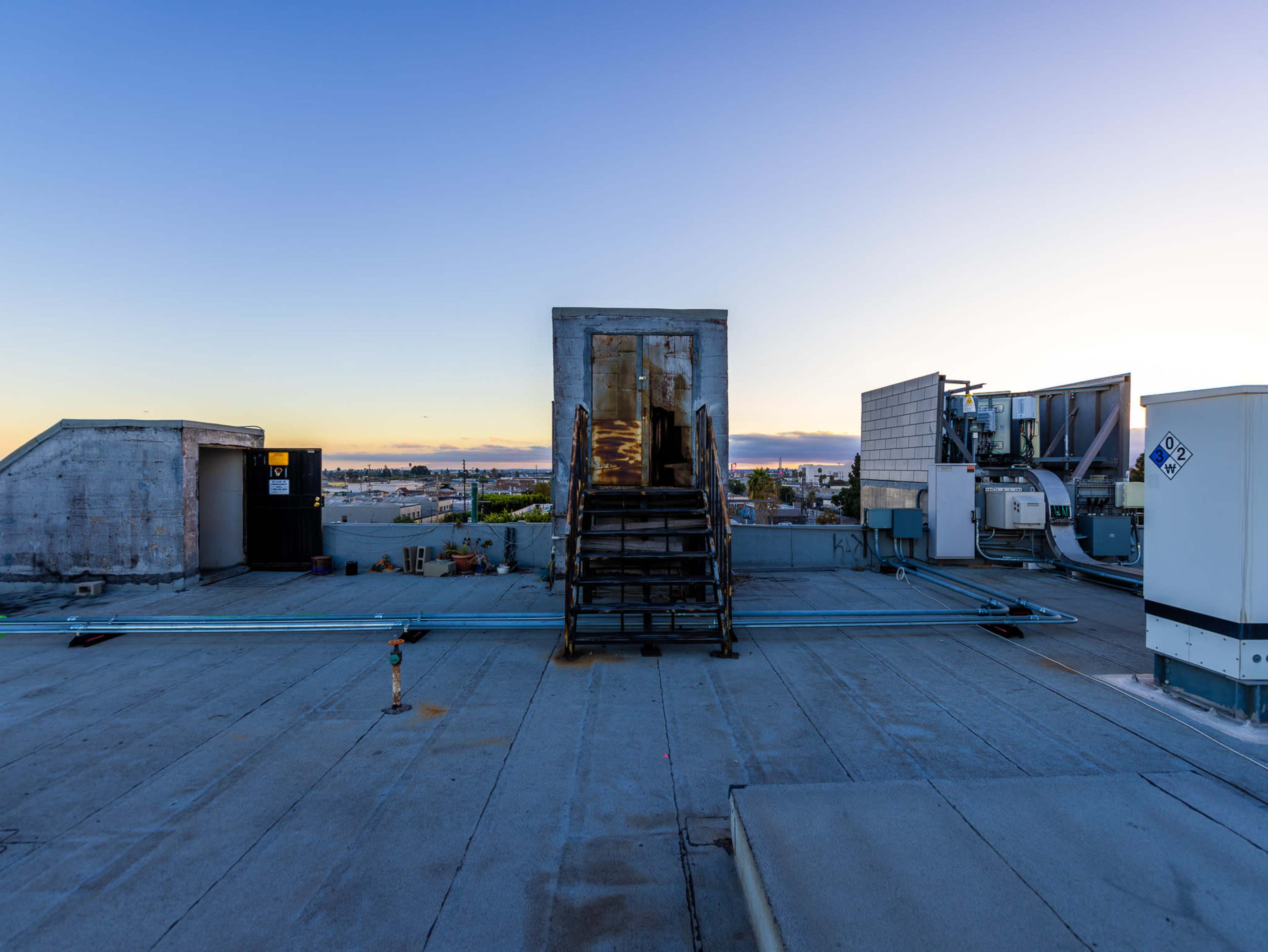 A rooftop with a metal staircase leading to a door, surrounded by air conditioning units and a clear sky at dusk.