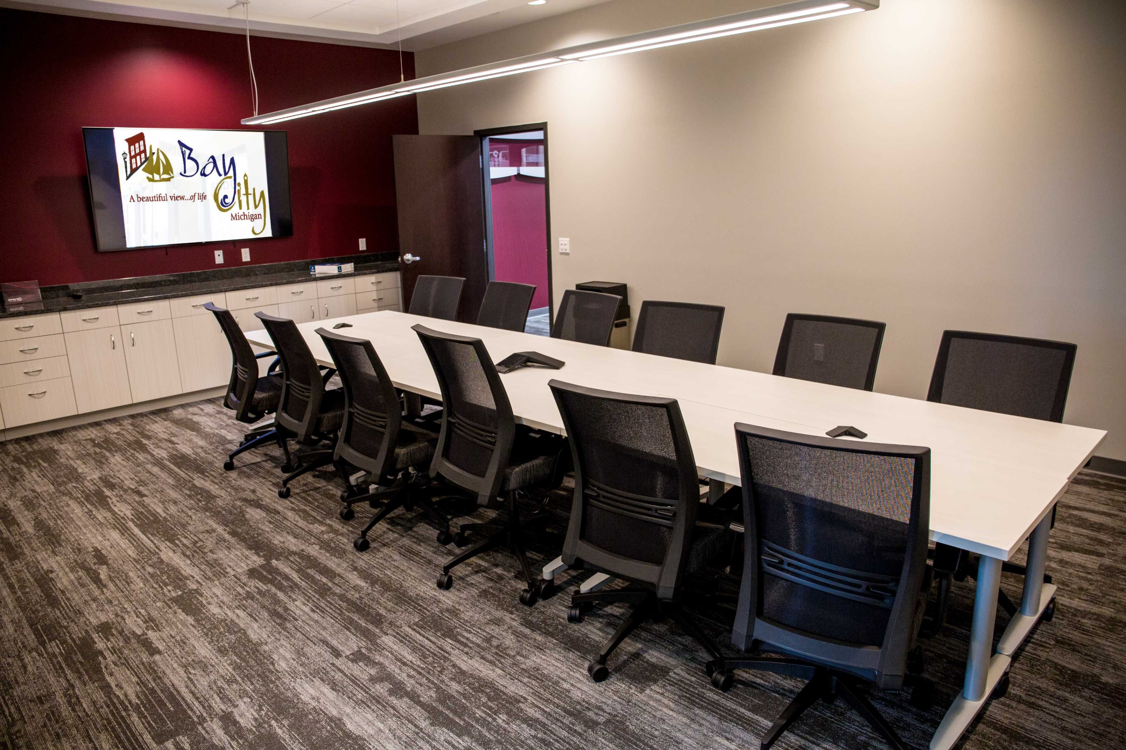 The image shows a modern conference room with a long white table, black ergonomic chairs, and a large wall-mounted screen displaying the text "Bay City."