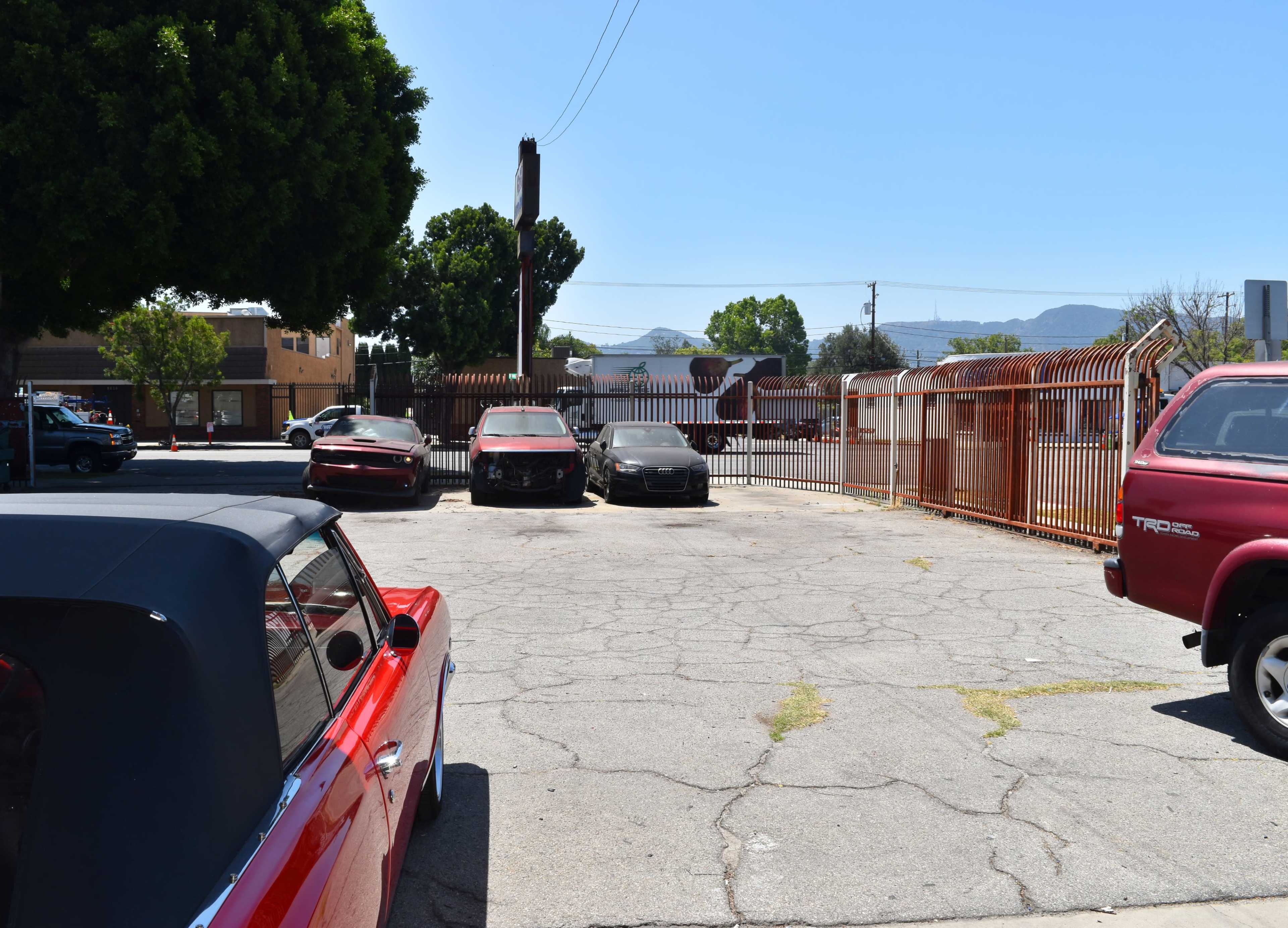 The image shows a parking lot with several cars, including a red convertible in the foreground, next to a fenced area and trees under a clear blue sky.