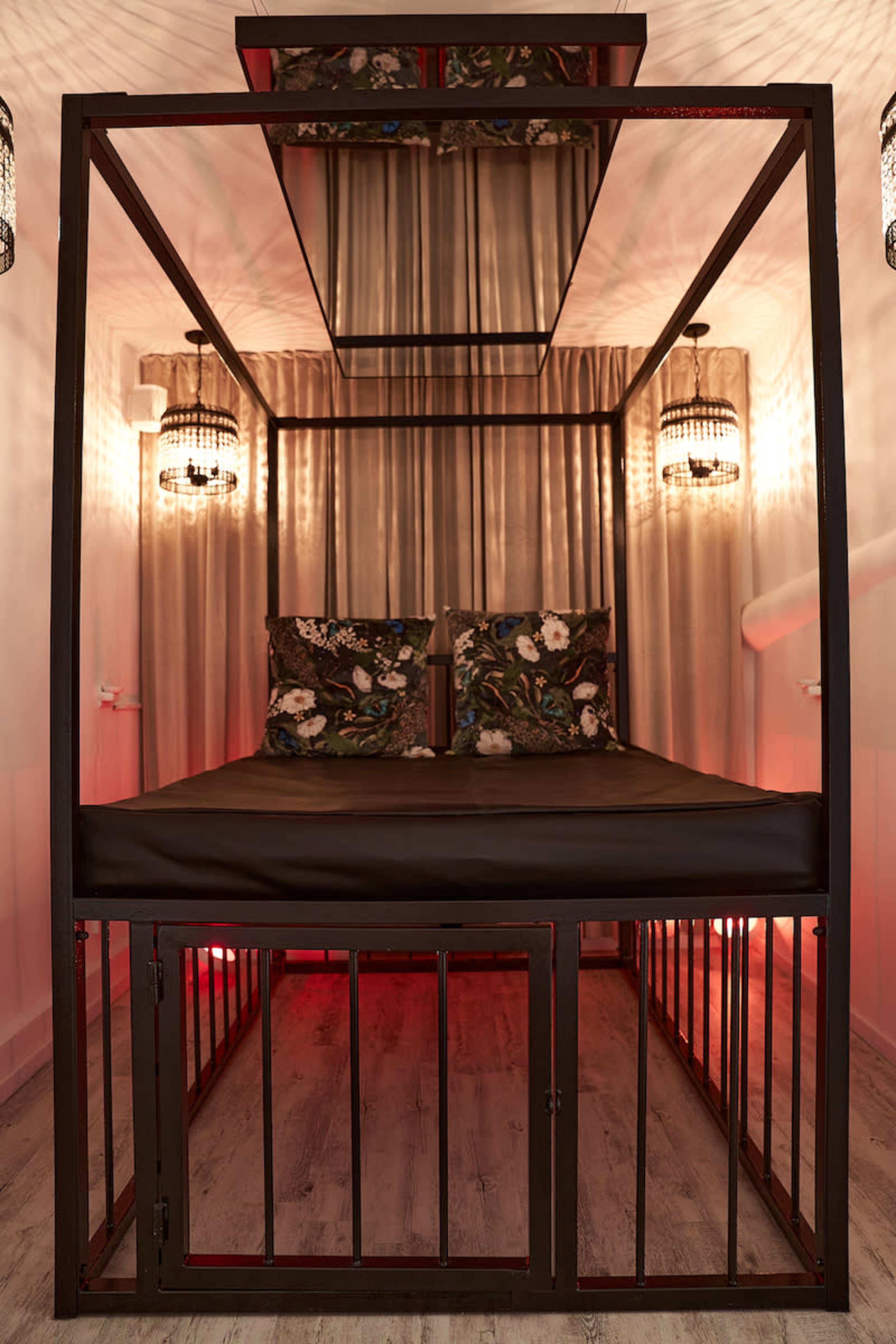 The image shows a metal-framed bed with decorative pillows, surrounded by curtains and dim lighting, resembling a cozy enclosure.