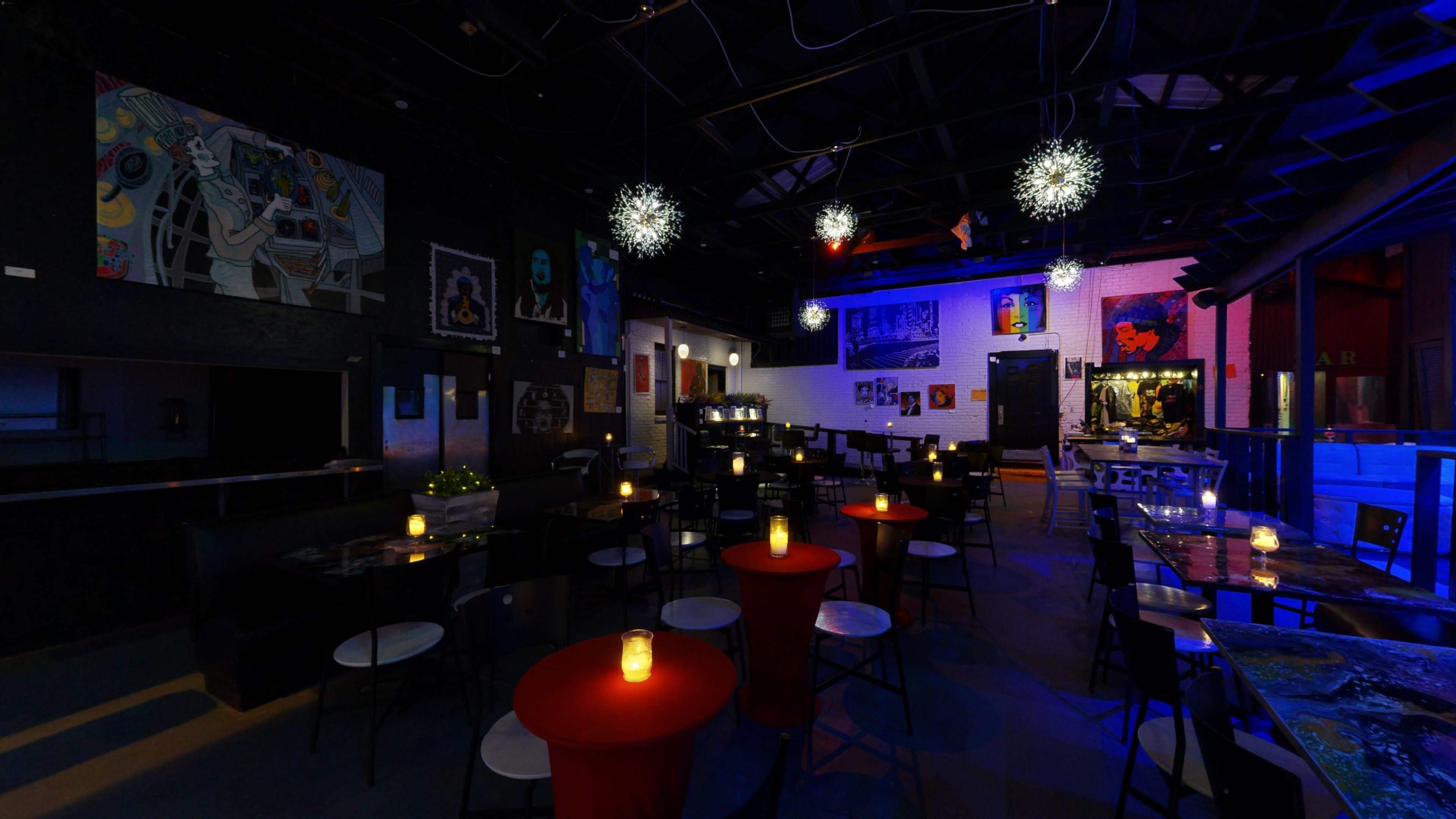 An indoor bar or restaurant with dim lighting, featuring art on the walls, tables with red cloths, and candles on each table.