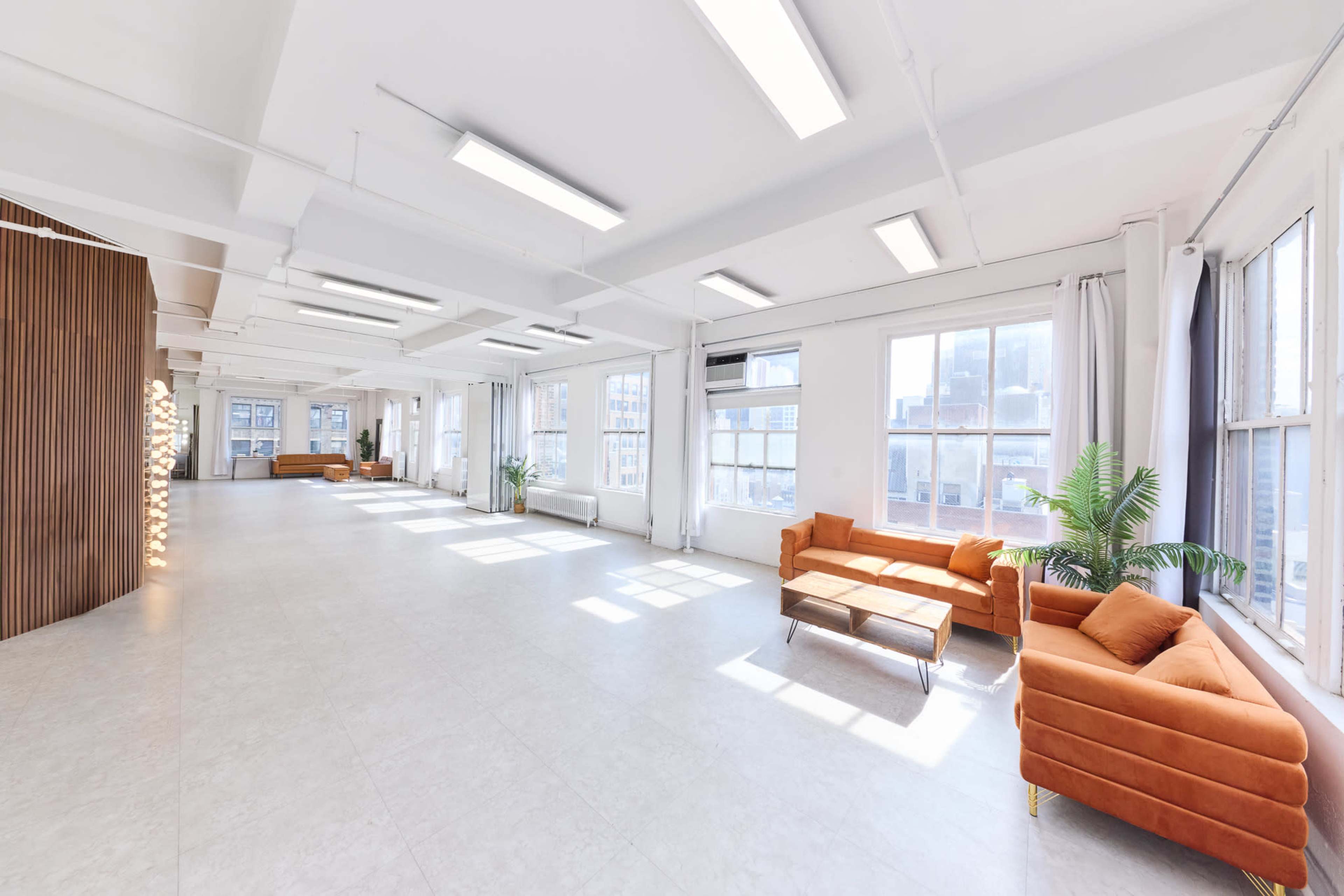 The image shows an empty, bright room with large windows, beige tiled flooring, and a few pieces of furniture, including an orange sofa and a wooden bench.