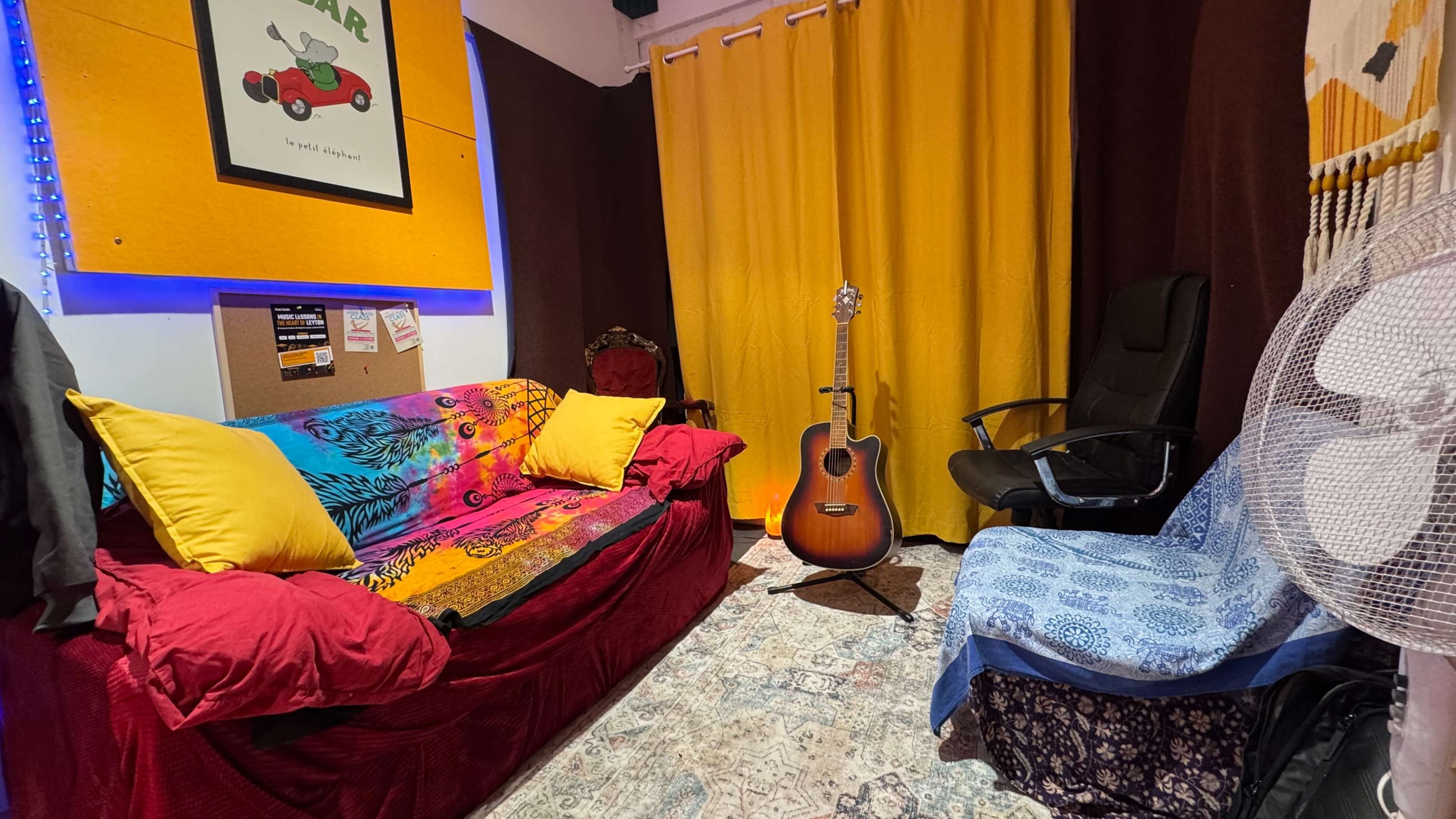 The image shows a cozy room with a red sofa covered with colorful blankets, a wooden acoustic guitar standing beside it, and a blue chair opposite, all set against yellow curtains.