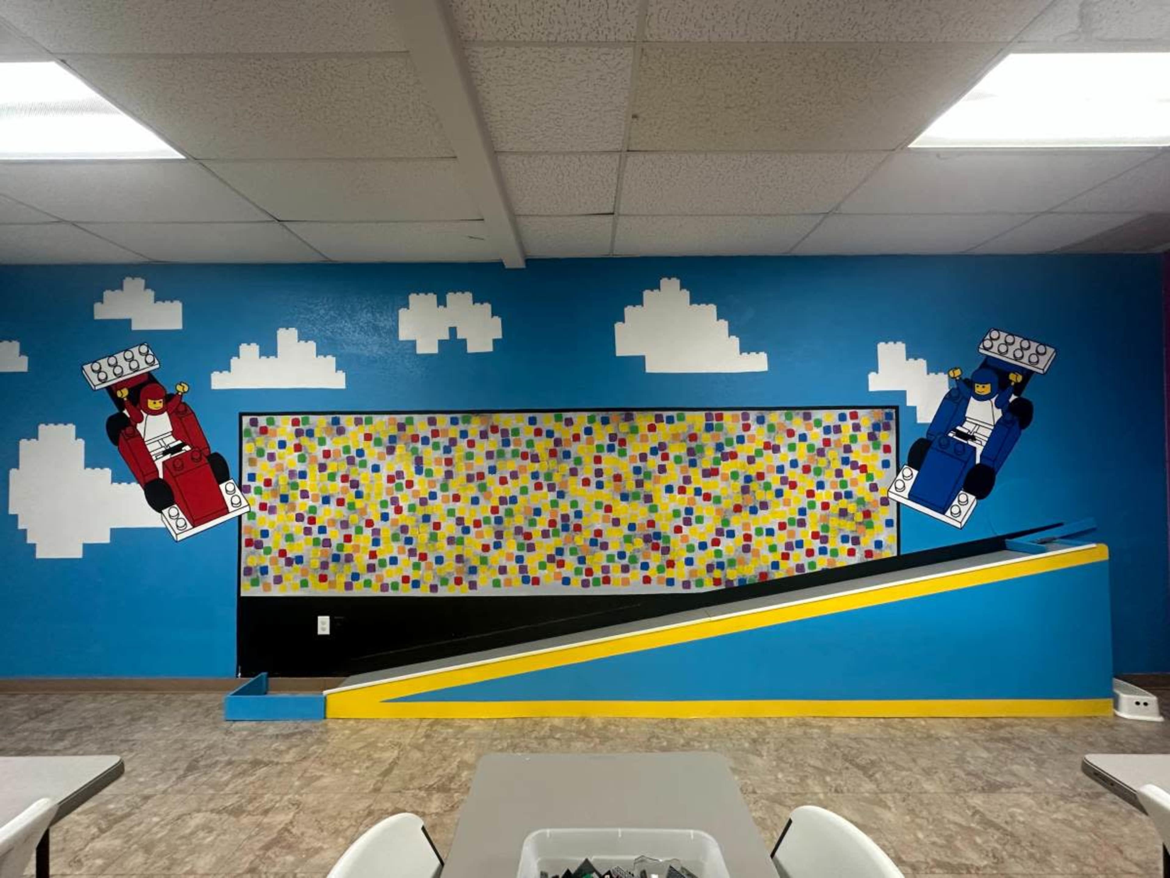 The image shows a colorful mural on a blue wall featuring two large LEGO-style figures, one in red and one in blue, alongside a stripe of yellow and a wall covered with multicolored sticky notes.