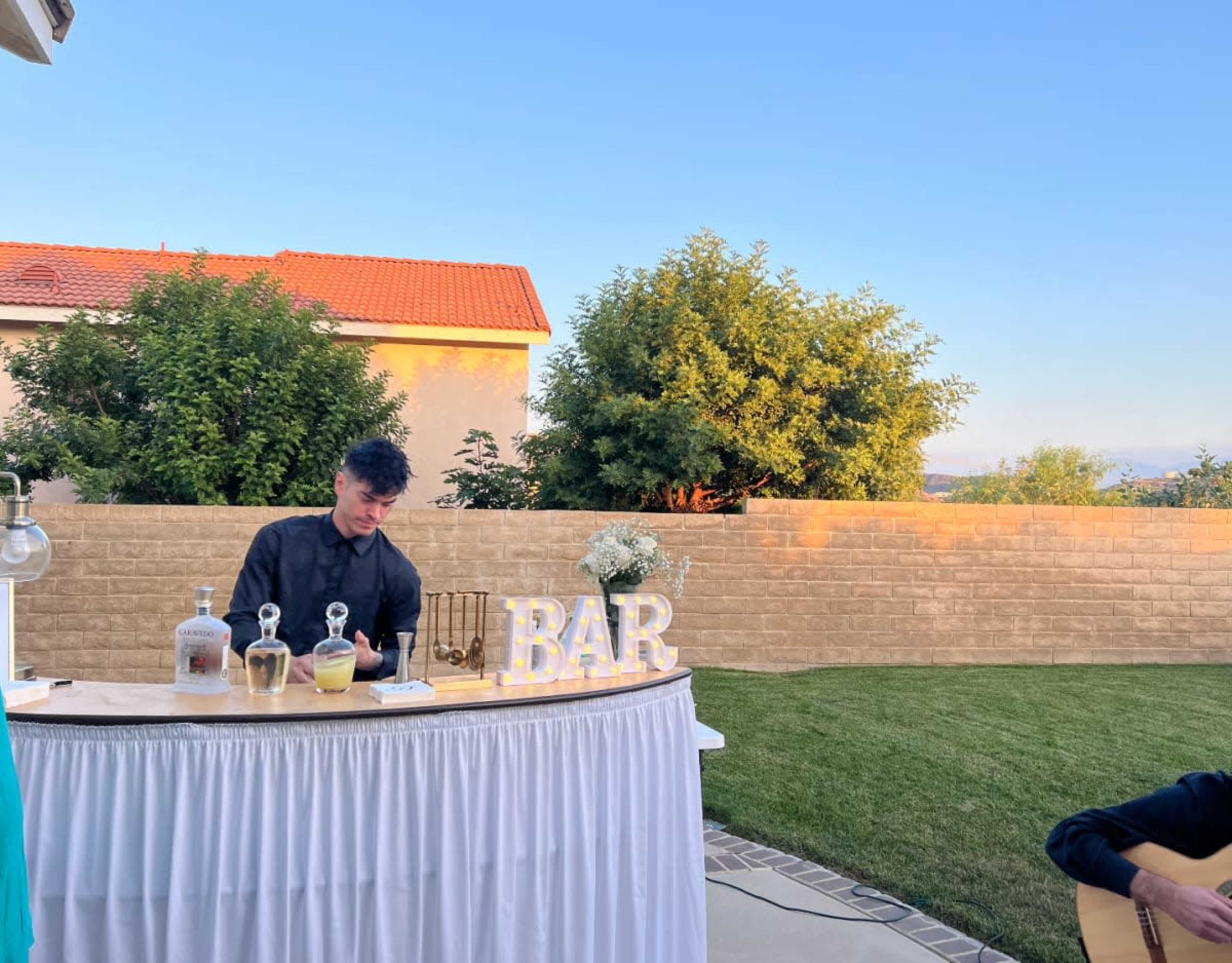 A bartender stands behind a wooden bar setup with drinks, while a person plays guitar nearby in a landscaped outdoor setting.