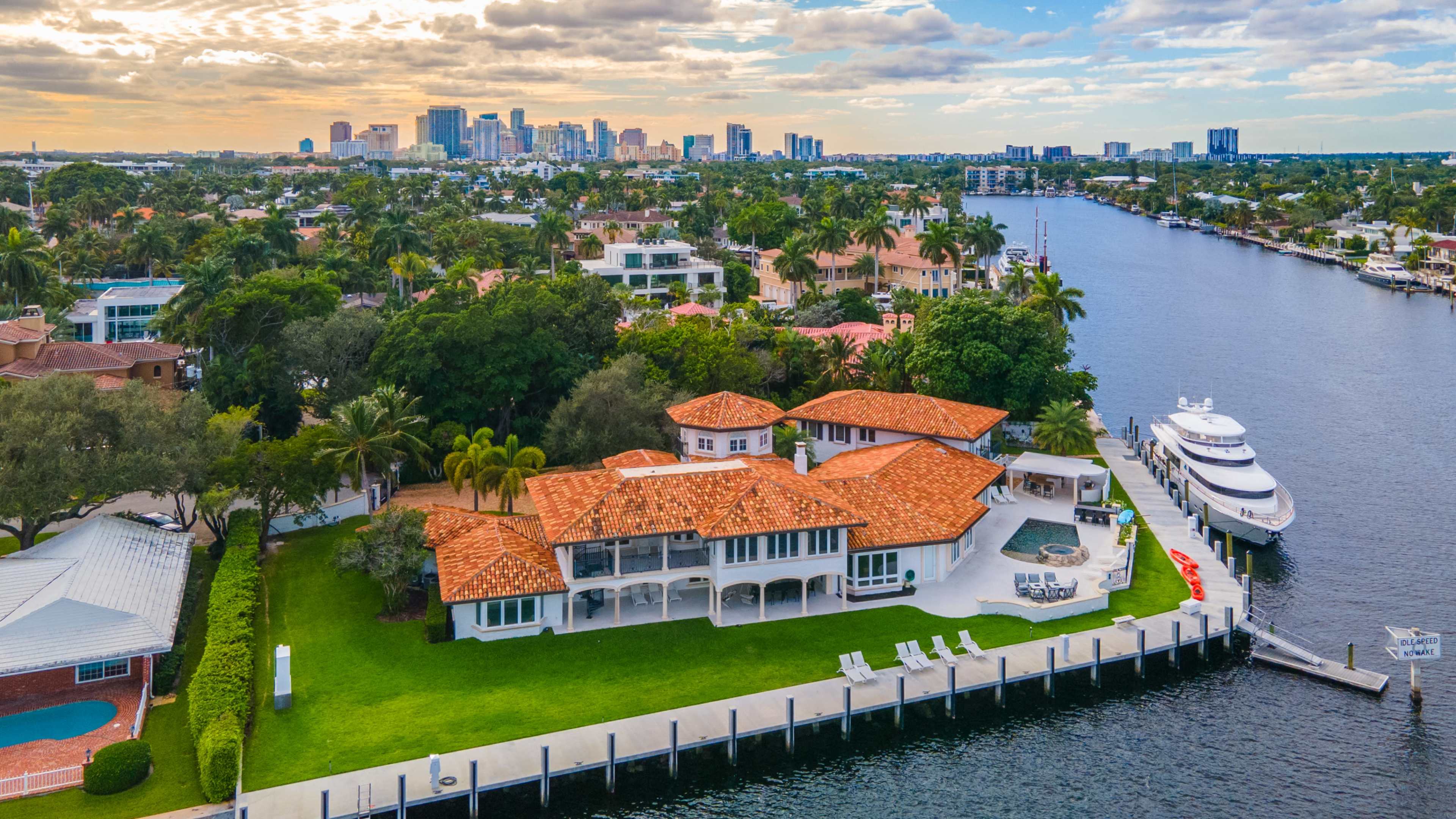 A waterfront mansion with a red-tiled roof is surrounded by lush greenery and overlooks a canal leading to a city skyline.