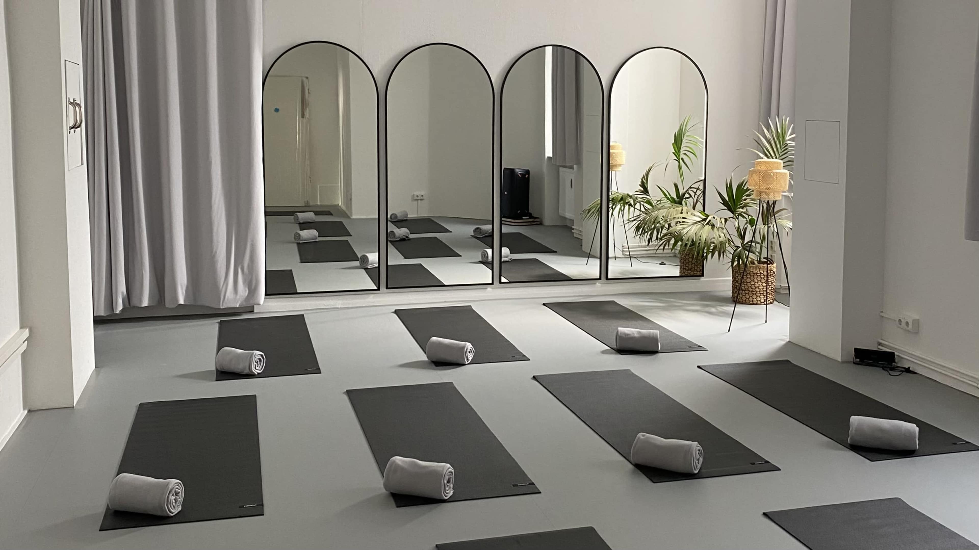 The image shows a minimalist yoga studio arranged with several yoga mats and rolled towels placed in front of three arched mirrors.