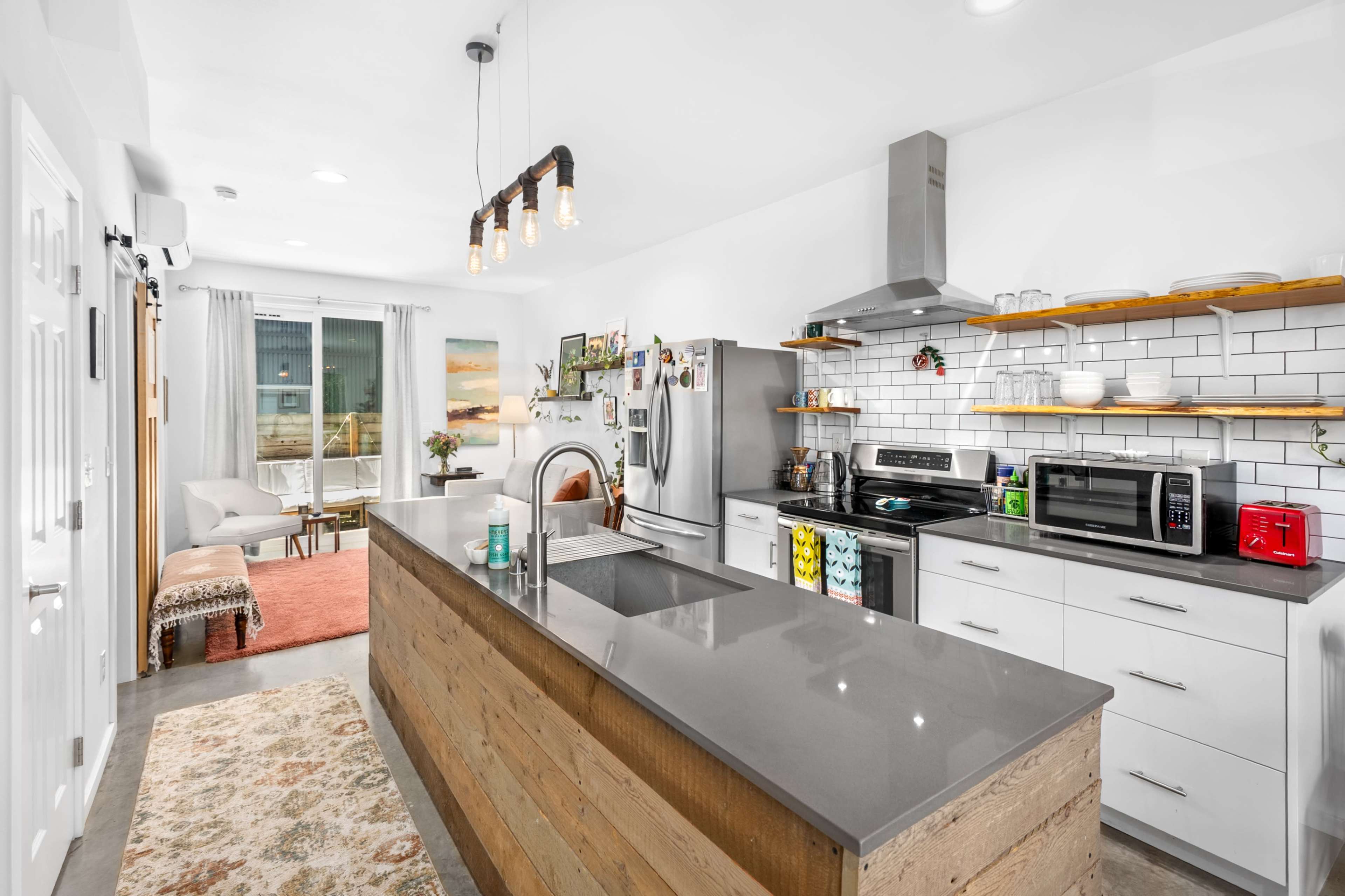 The image shows a modern kitchen with a grey island, wooden accents, and stainless steel appliances, featuring an open layout that leads to a living area with a sofa and large windows.