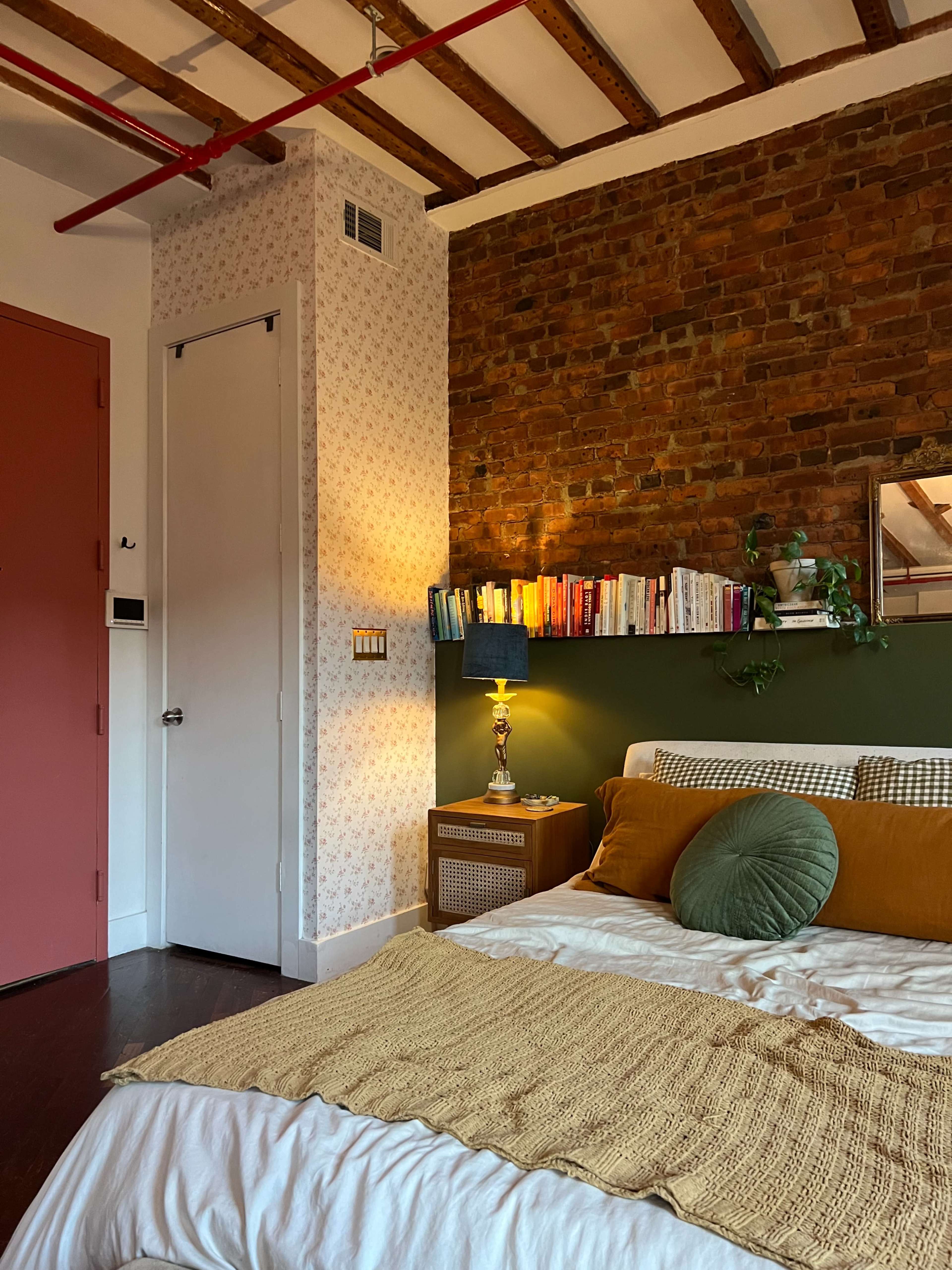 A cozy bedroom features a brick accent wall, a bed with a green headboard, and a bookshelf above a nightstand.