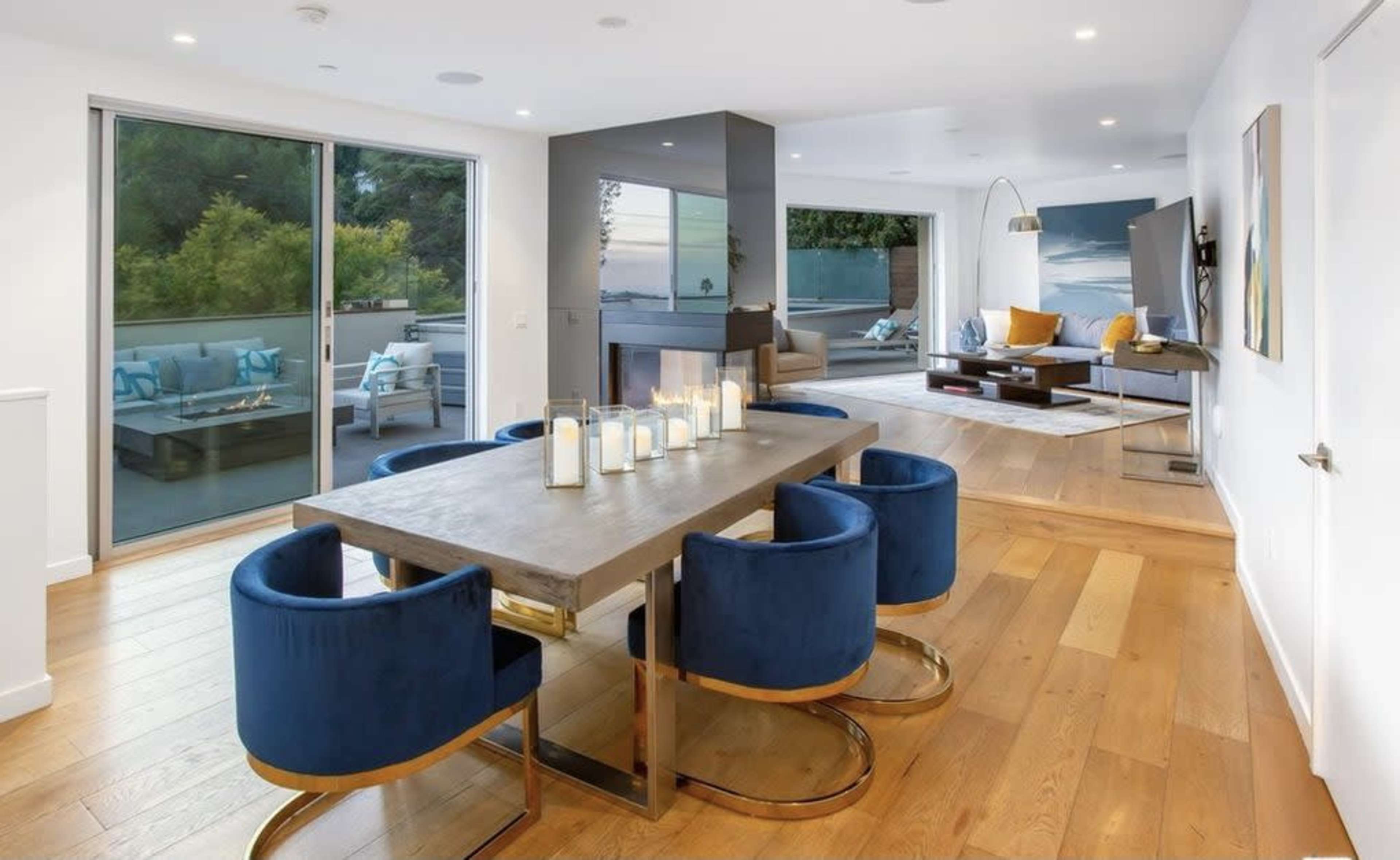 The image shows a modern living space featuring a dining area with a rectangular table surrounded by blue upholstered chairs, a sleek gray partition, and a cozy lounge area with a view of an outdoor terrace.