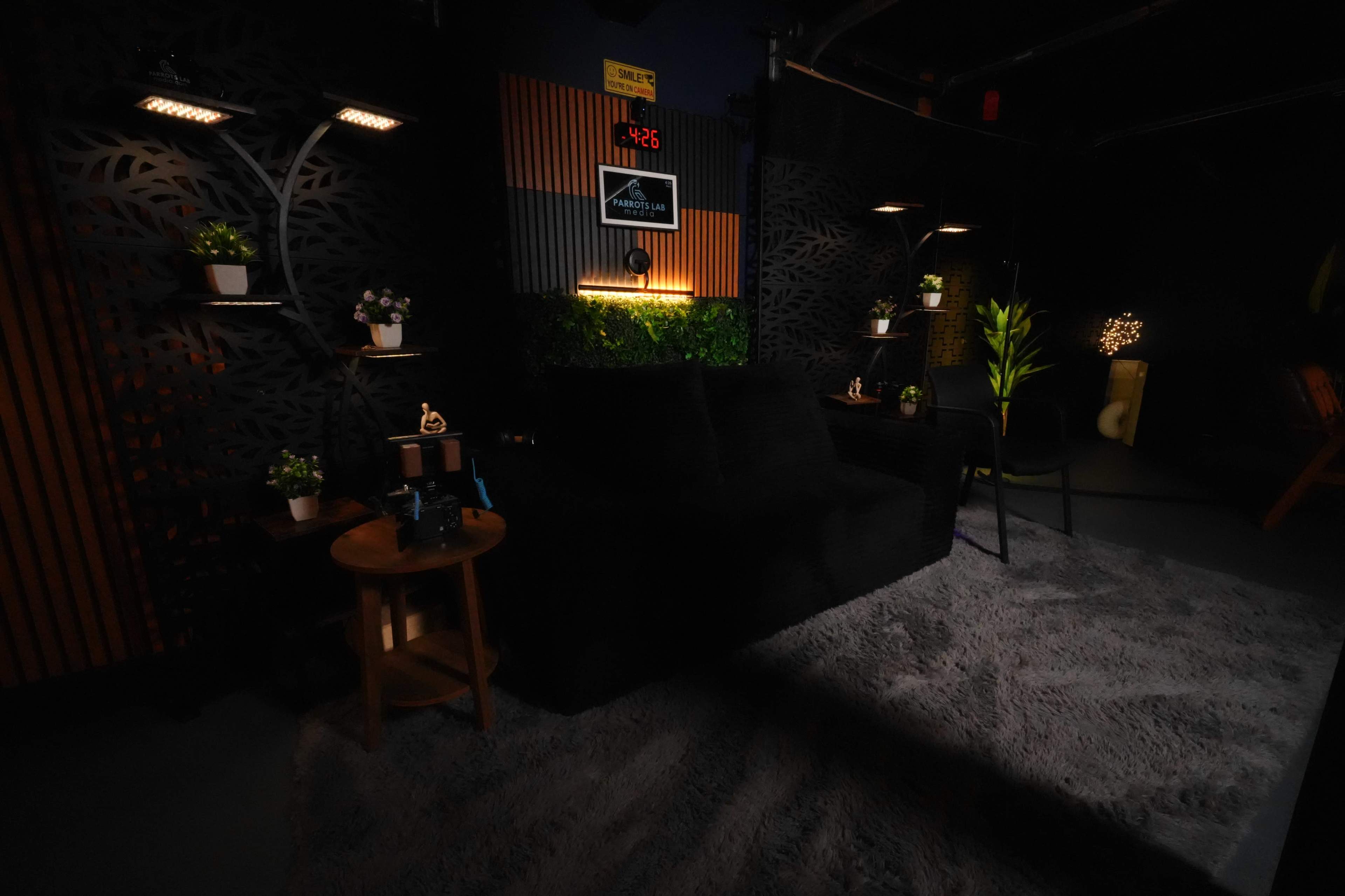 The image shows a dark, modern lounge area with a large couch, shelves with potted plants, and ambient lighting.