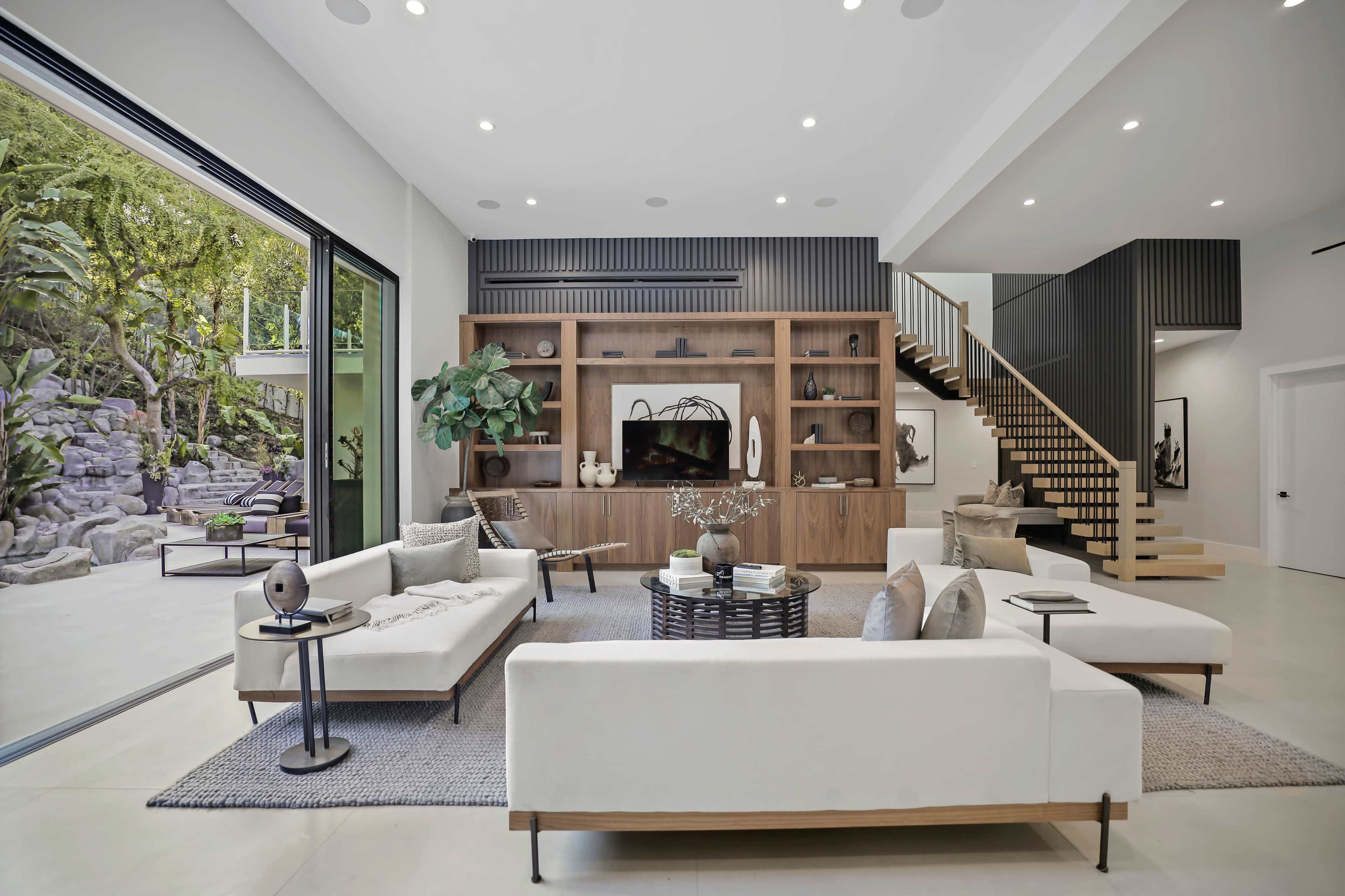 The image shows a modern living room with a large seating area, a wooden media console, and a staircase leading to an upper level, beside a glass wall that opens to an outdoor space.