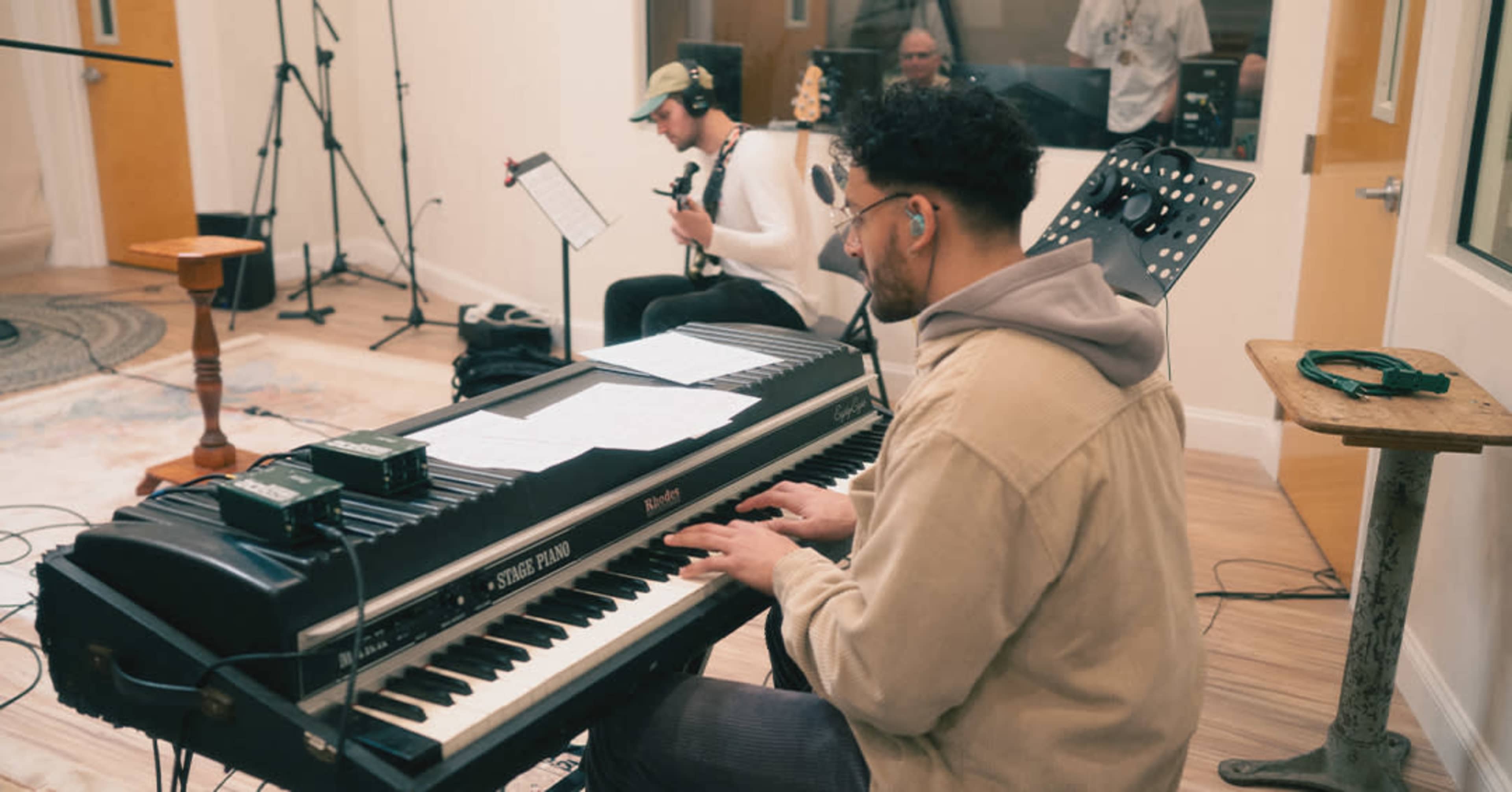 A musician plays a keyboard in a recording studio while another musician sits in the background with a guitar.
