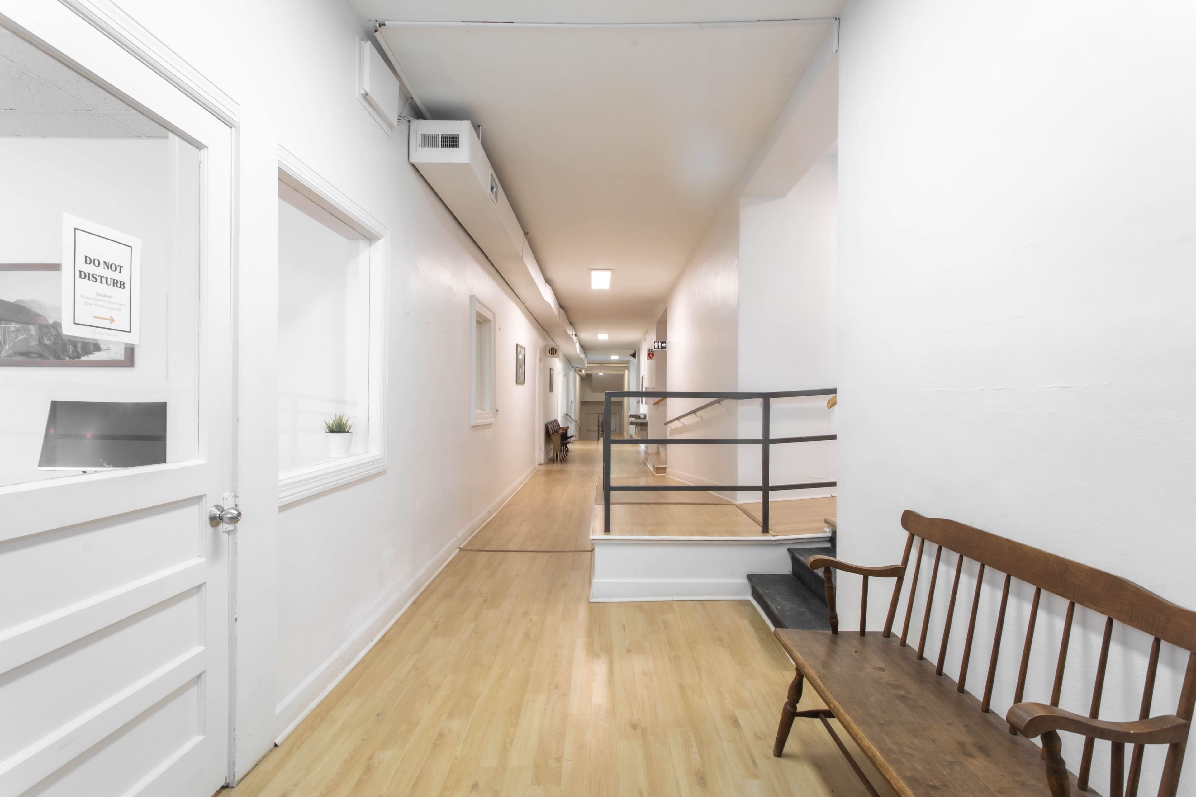 The image shows a long hallway with wooden flooring, a bench on one side, and doors leading to other rooms.