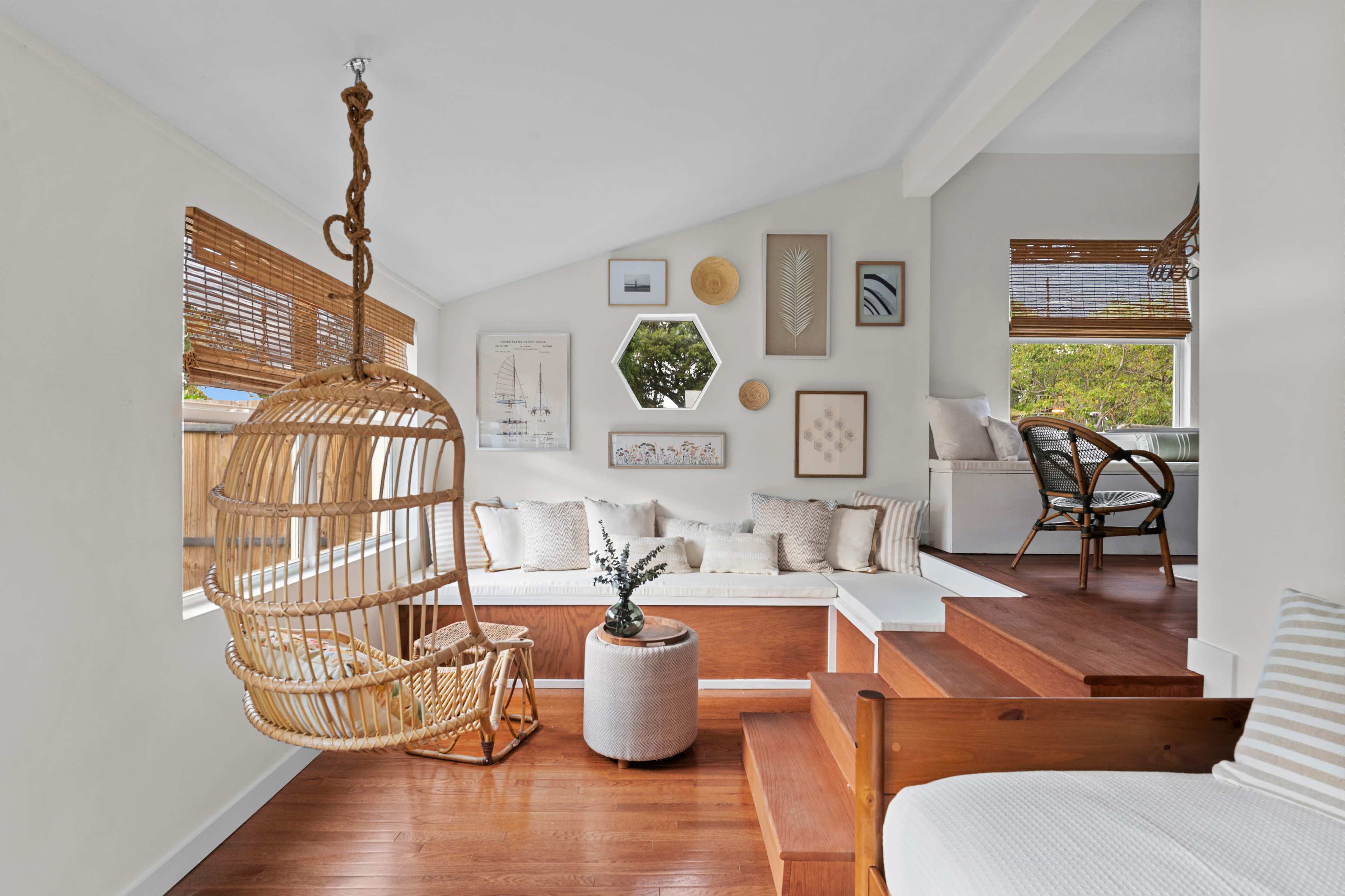A cozy living space features a hanging chair, a built-in bench with decorative pillows, and art on the walls, complemented by wooden accents and large windows.