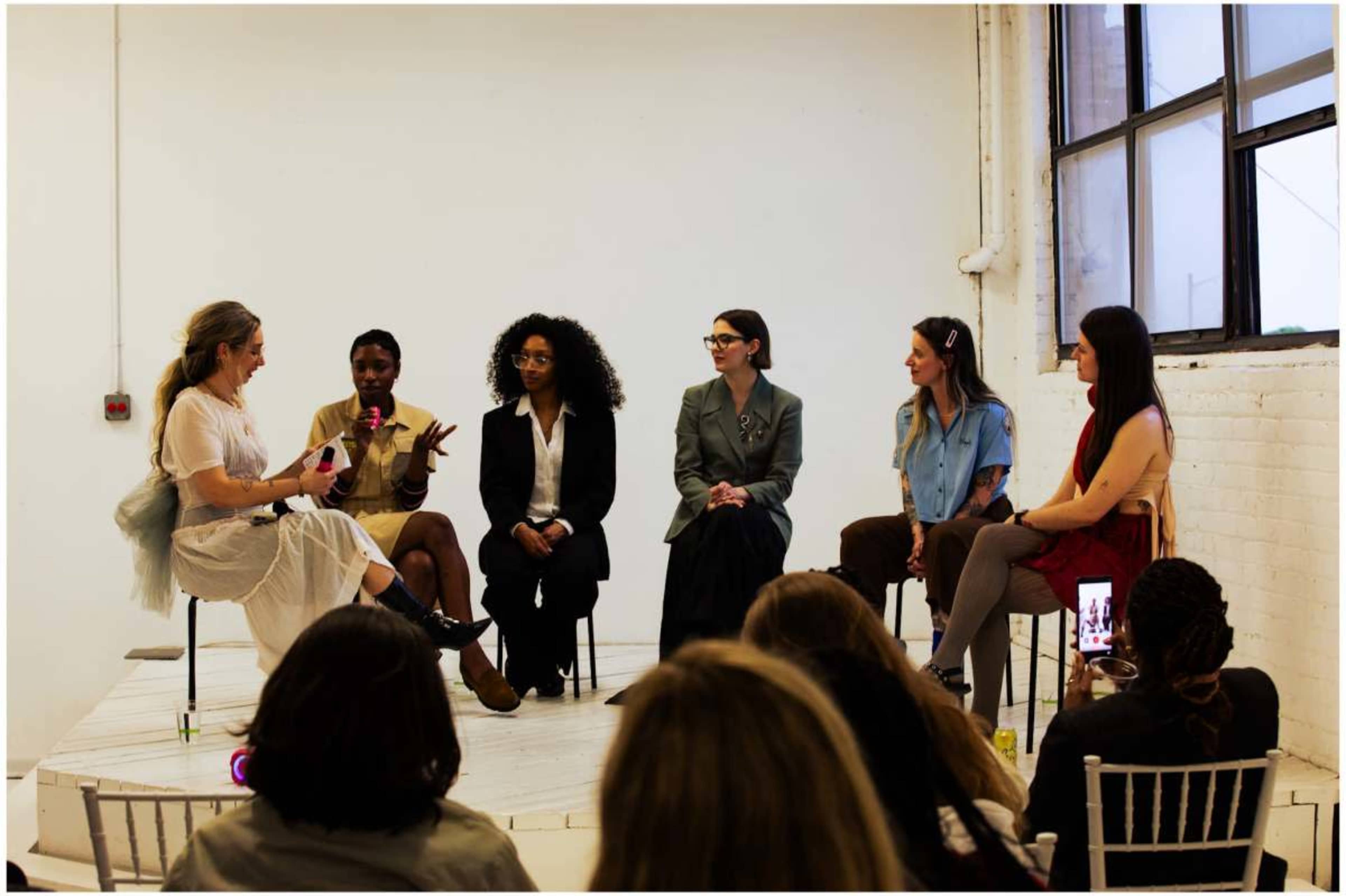 A group of five women sit in a semi-circle on a raised platform, engaged in discussion while an audience watches.