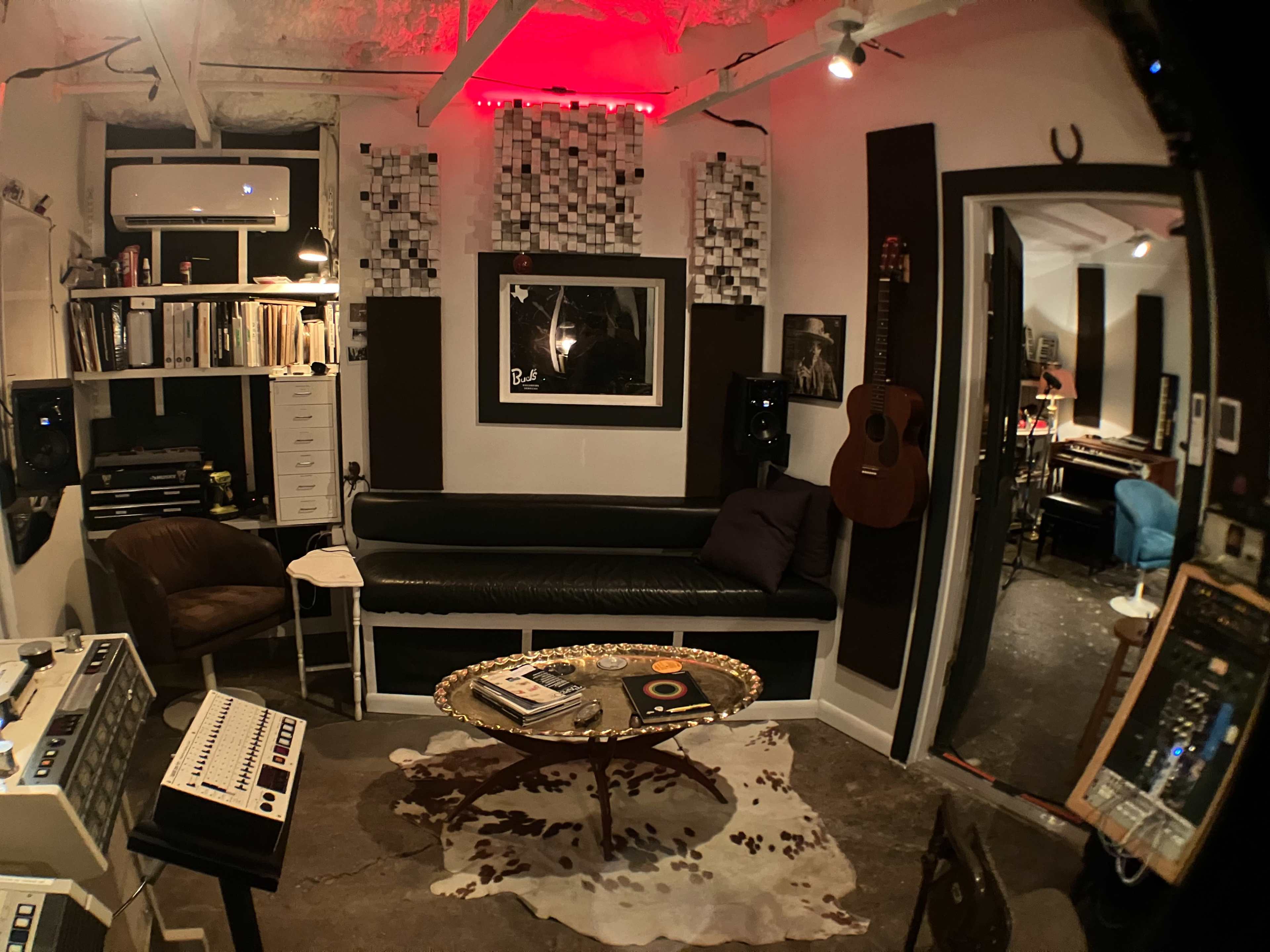 The image depicts a cozy recording studio featuring a leather couch, a coffee table with records, and various musical equipment surrounded by art and acoustic panels on the walls.
