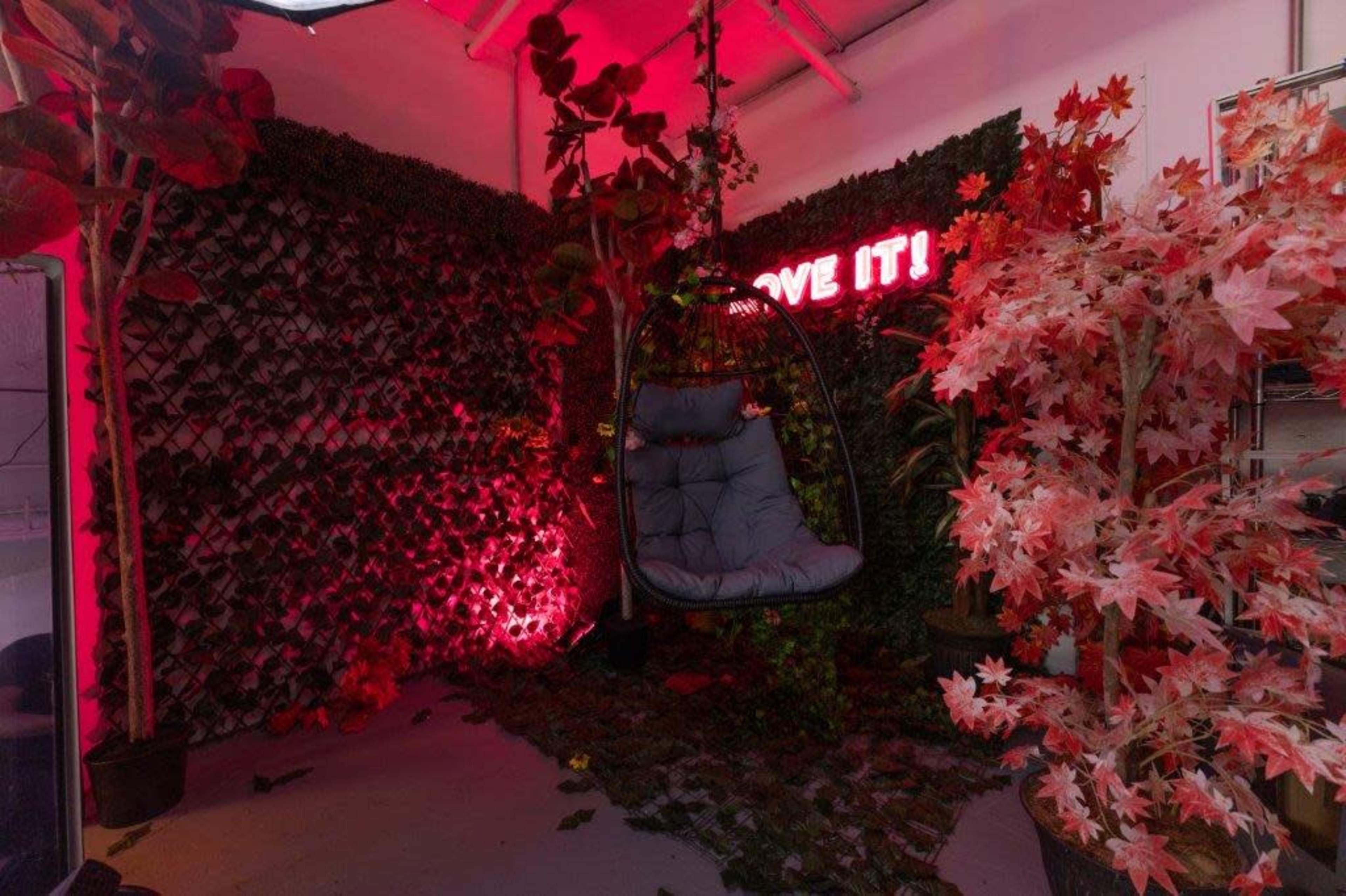 A cozy indoor space with a swing chair surrounded by green plants and a neon sign that reads "LOVE IT!" illuminated in red light.