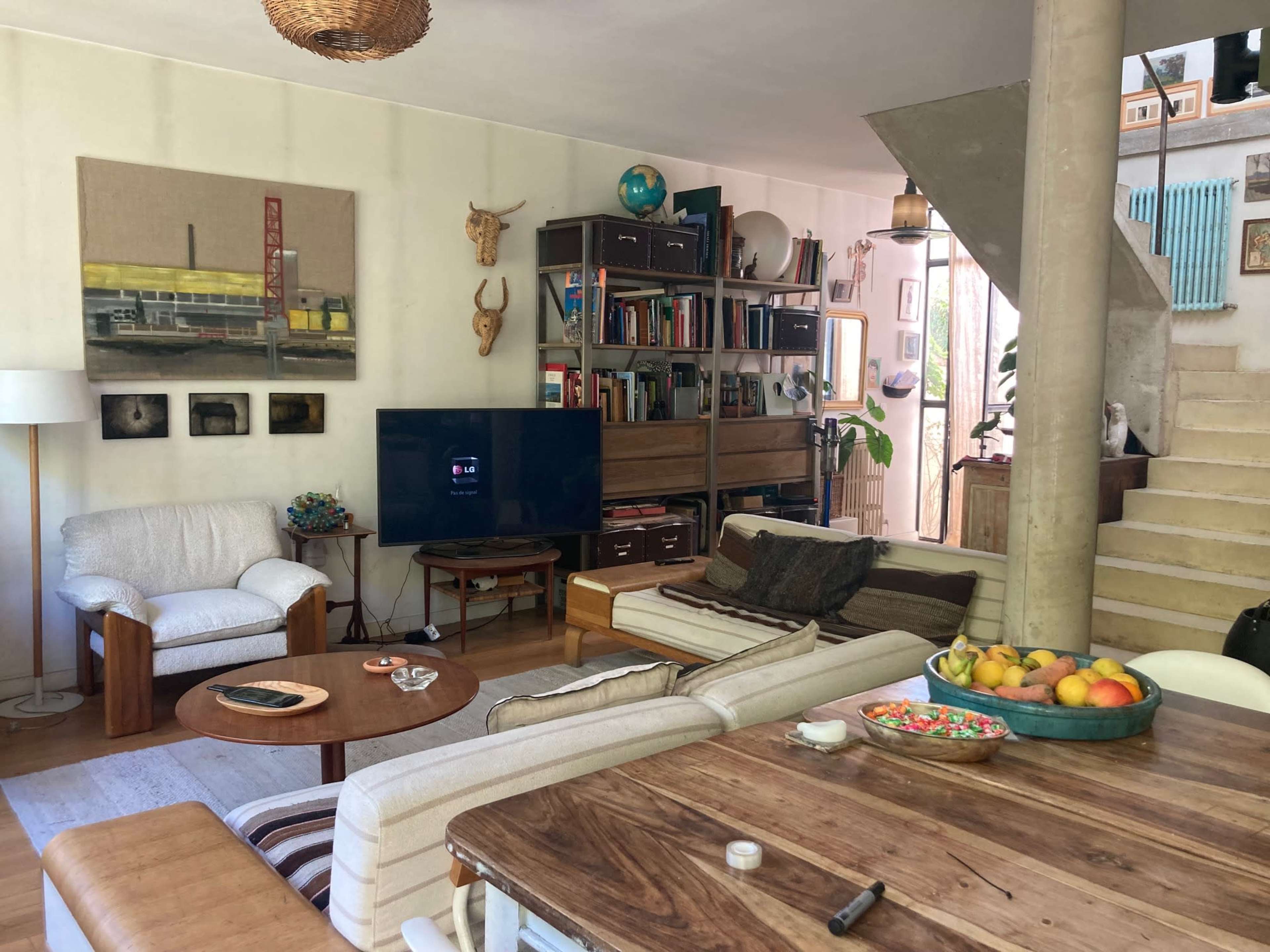 The image shows a cozy living room with a mix of wooden furniture, a large TV, bookshelves, and a staircase leading to an upper level.
