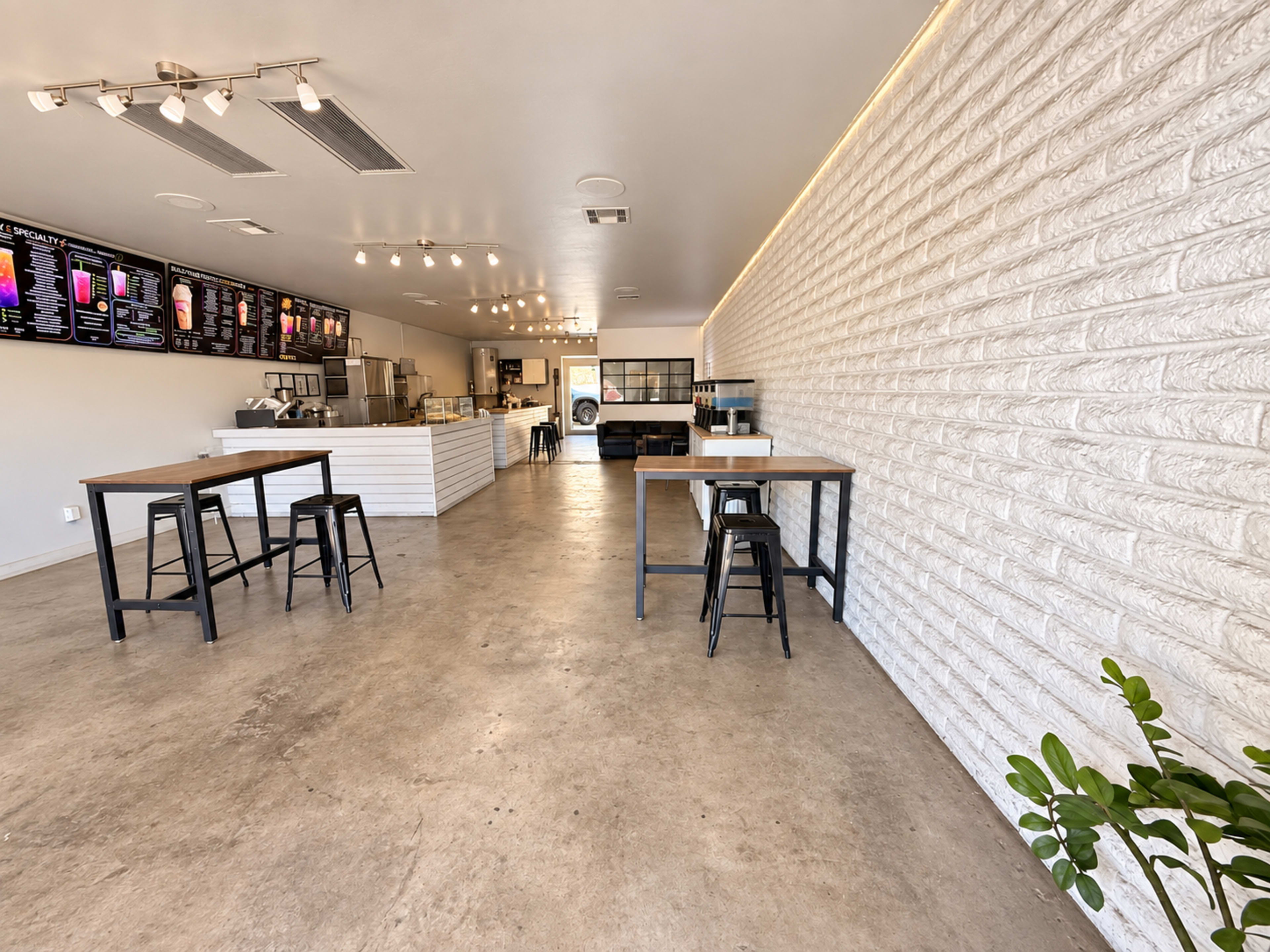 The image shows a modern, minimalist café interior featuring white walls, concrete flooring, and several high tables with black stools.