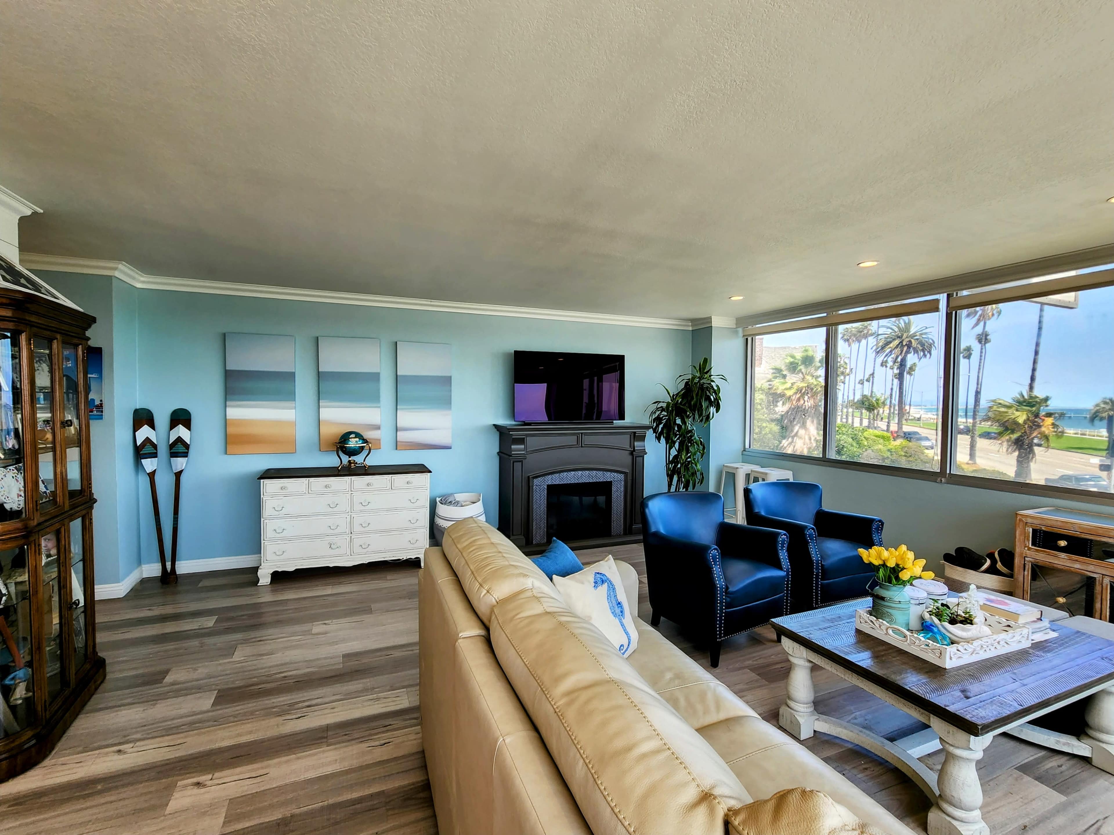 The living room features a light-colored sofa, two blue chairs, a fireplace, a television, and large windows with a view of palm trees and the beach.