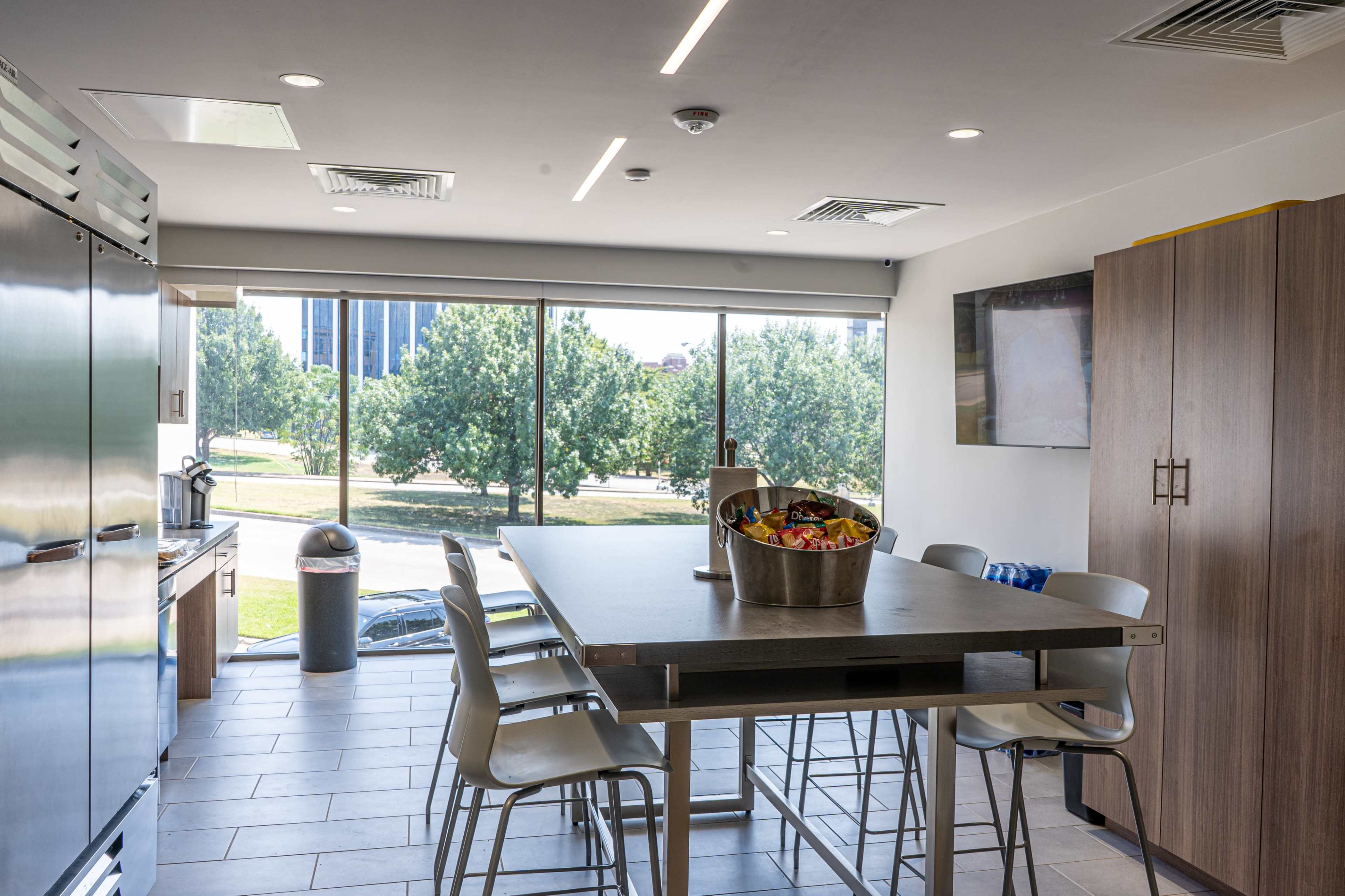 A modern kitchen features a large table with metal chairs, stainless steel appliances, and large windows showcasing a view of trees outside.
