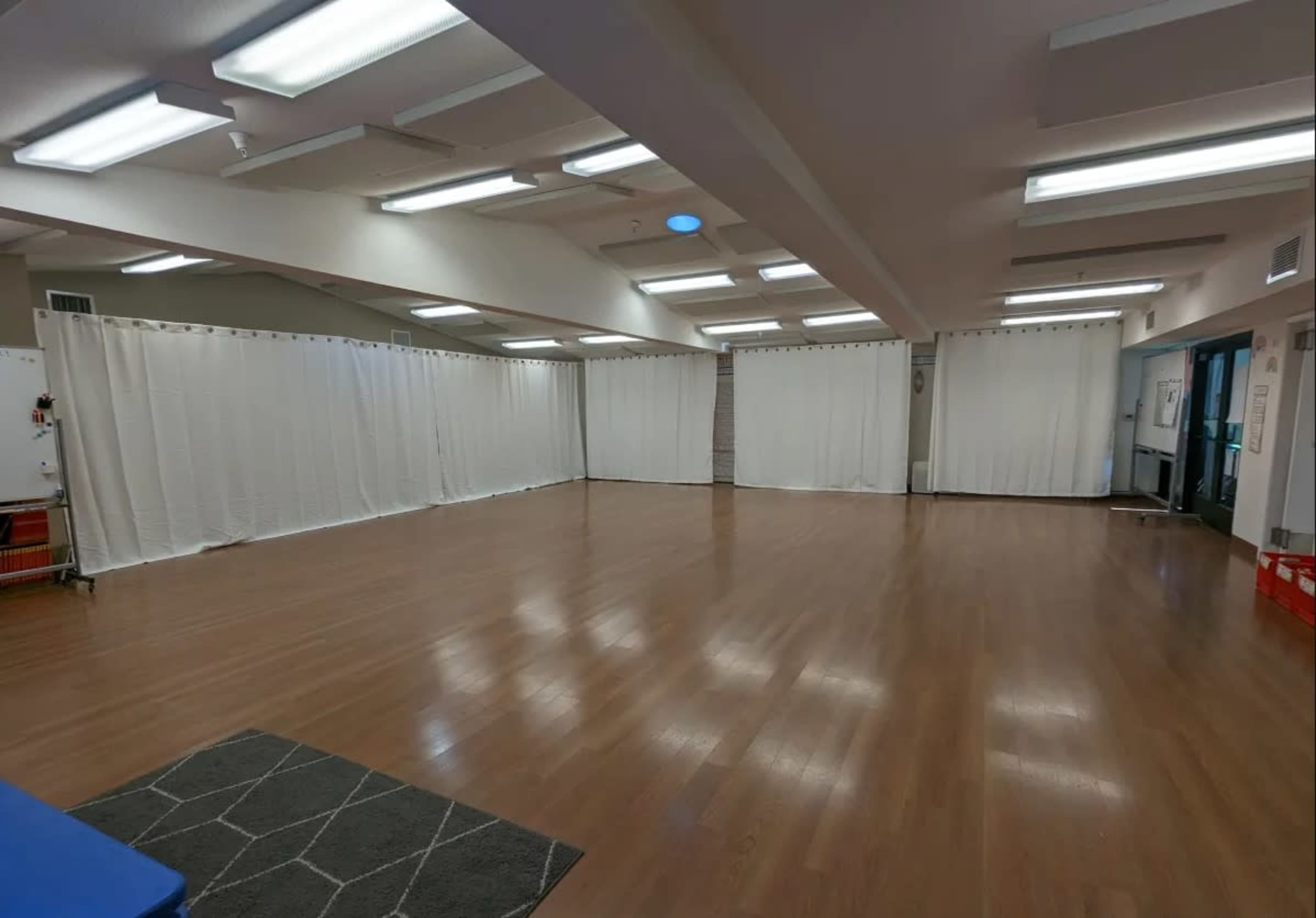 The image shows a spacious room with wooden flooring, white curtains dividing the space, and bright overhead lighting.