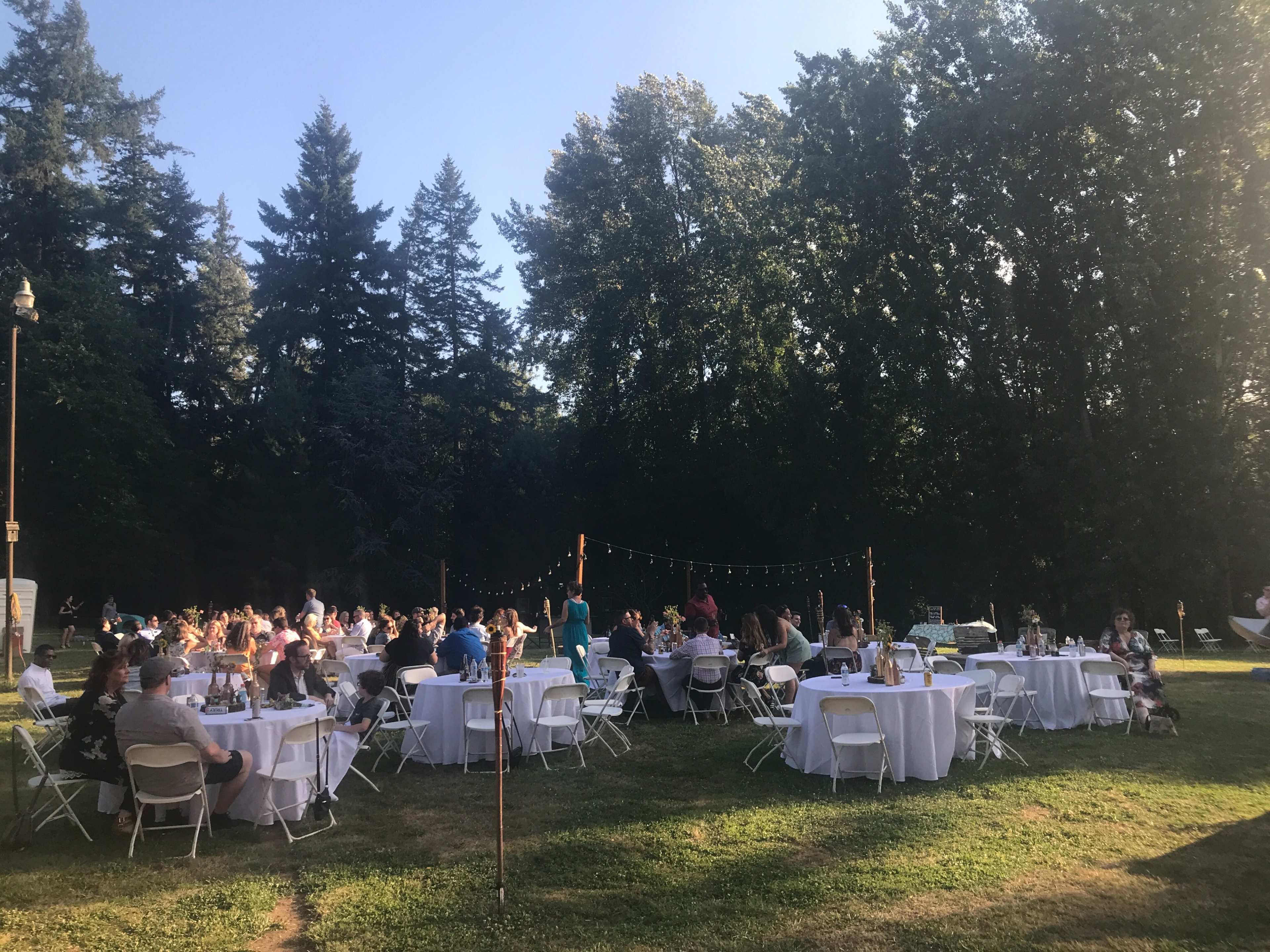 A large outdoor gathering is taking place with numerous round tables set up under string lights amidst trees.