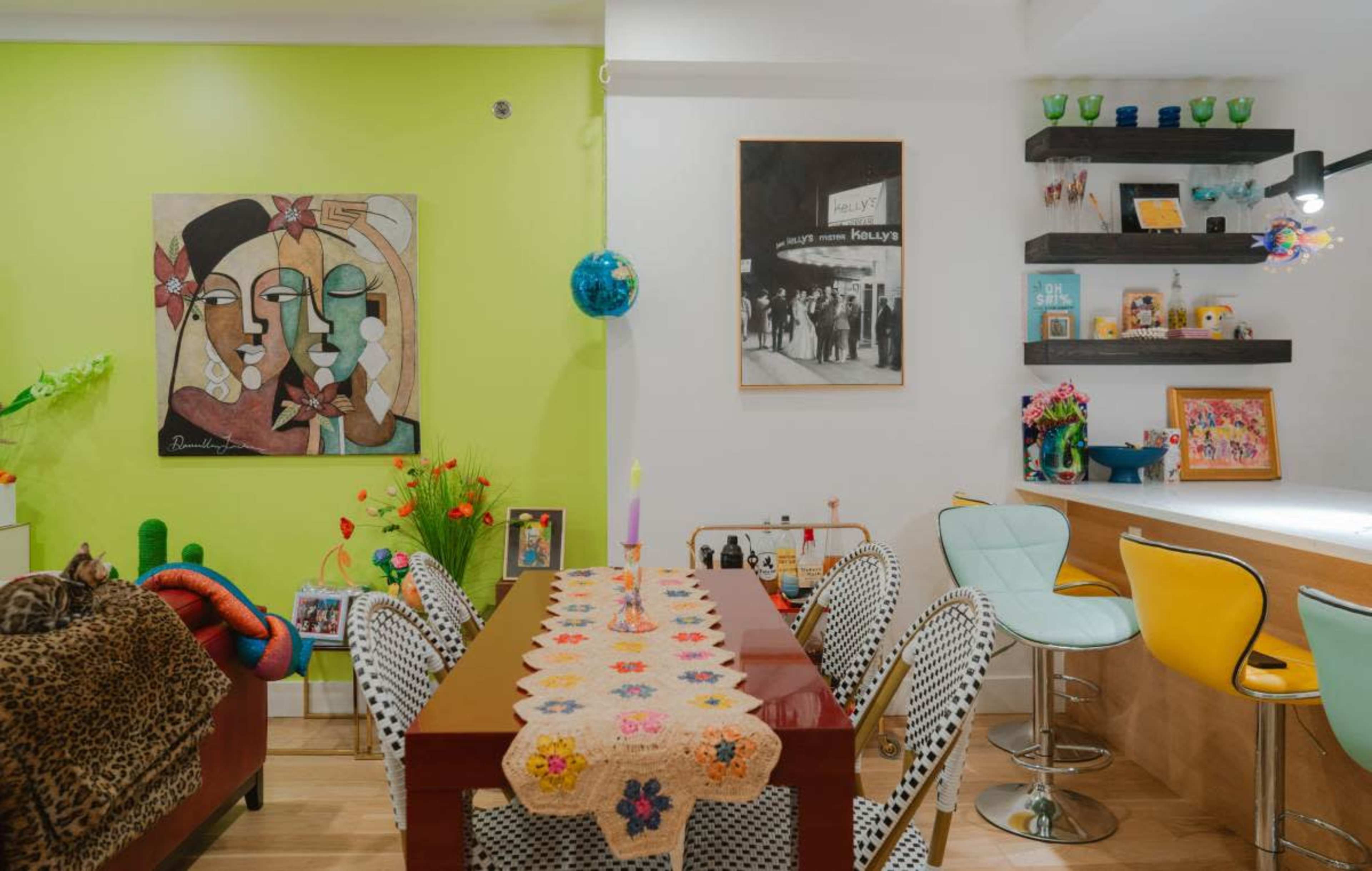 A colorful dining area with a table covered in a crocheted tablecloth, surrounded by decorative chairs, and a wall adorned with artwork and shelves displaying various items.